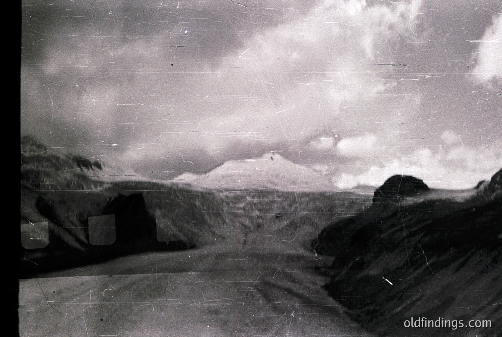 Vintage black-and-white alpine landscape with rugged terrain and distant snow-capped peak. Foreground shows weathered stone walls, likely from a historic farm or village. Dramatic cloud formations suggest early 20th-century photographic techniques.