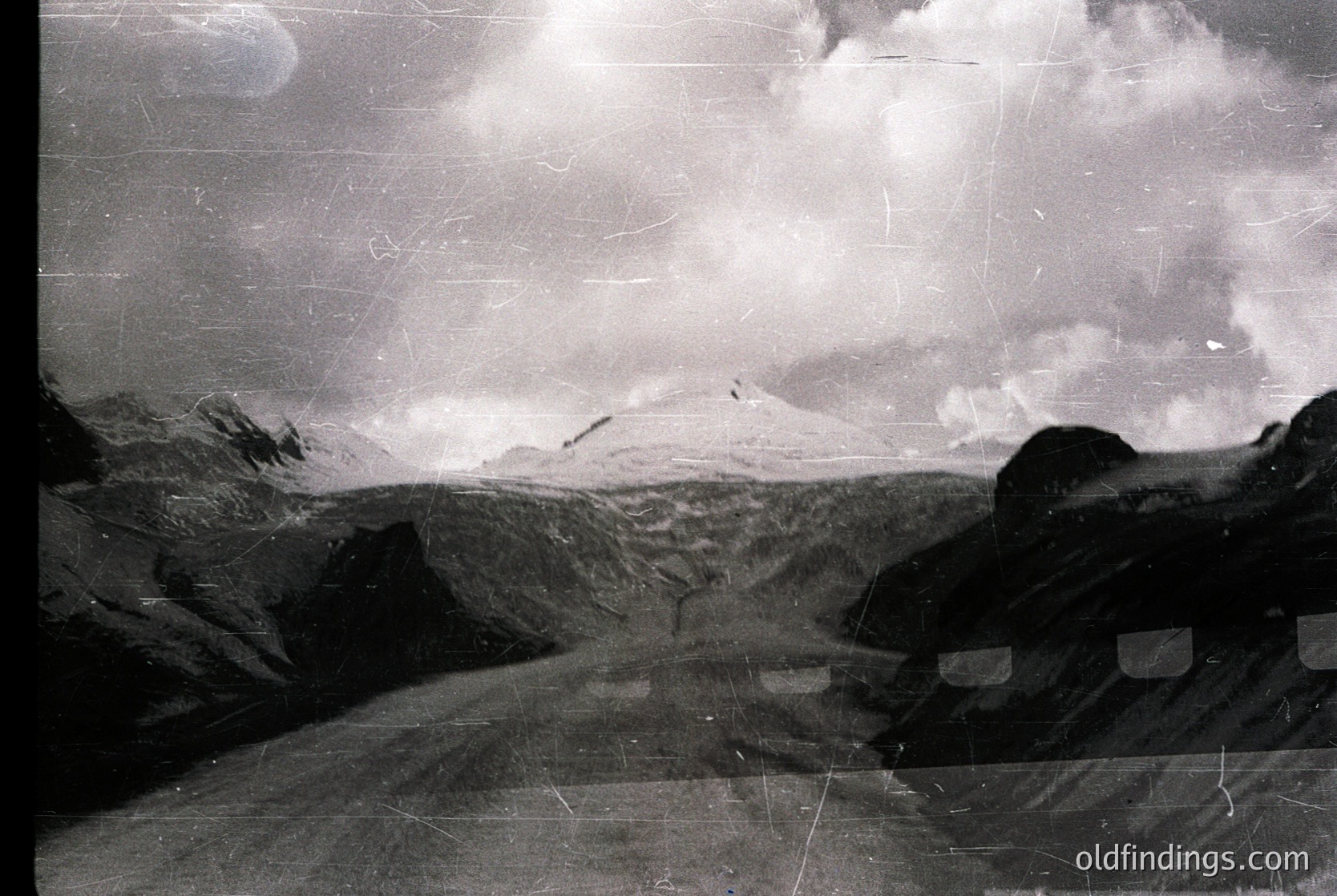 Vintage black-and-white alpine landscape with rugged peaks and dramatic cloud formations. Foreground shows weathered stone steps or path, suggesting a hiking trail. Texture indicates aged film grain and potential scratches. Likely mid-20th century mountaineering or travel documentation.