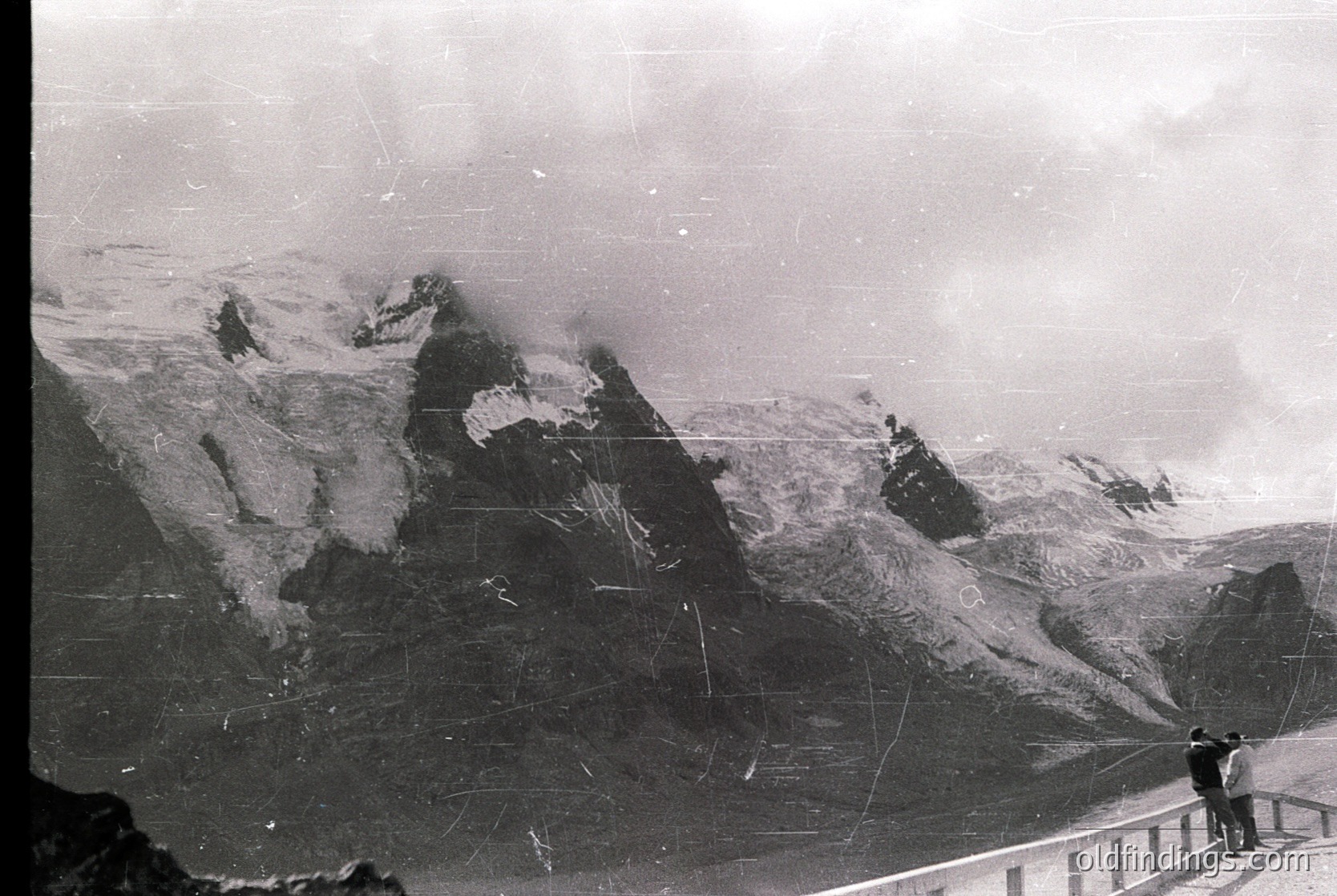 Vintage black-and-white alpine scene featuring jagged peaks and snow-covered slopes. A lone figure stands on a viewing platform, framed by a railing. Likely early-to-mid 20th century, suggesting historical tourism or mountaineering interest.