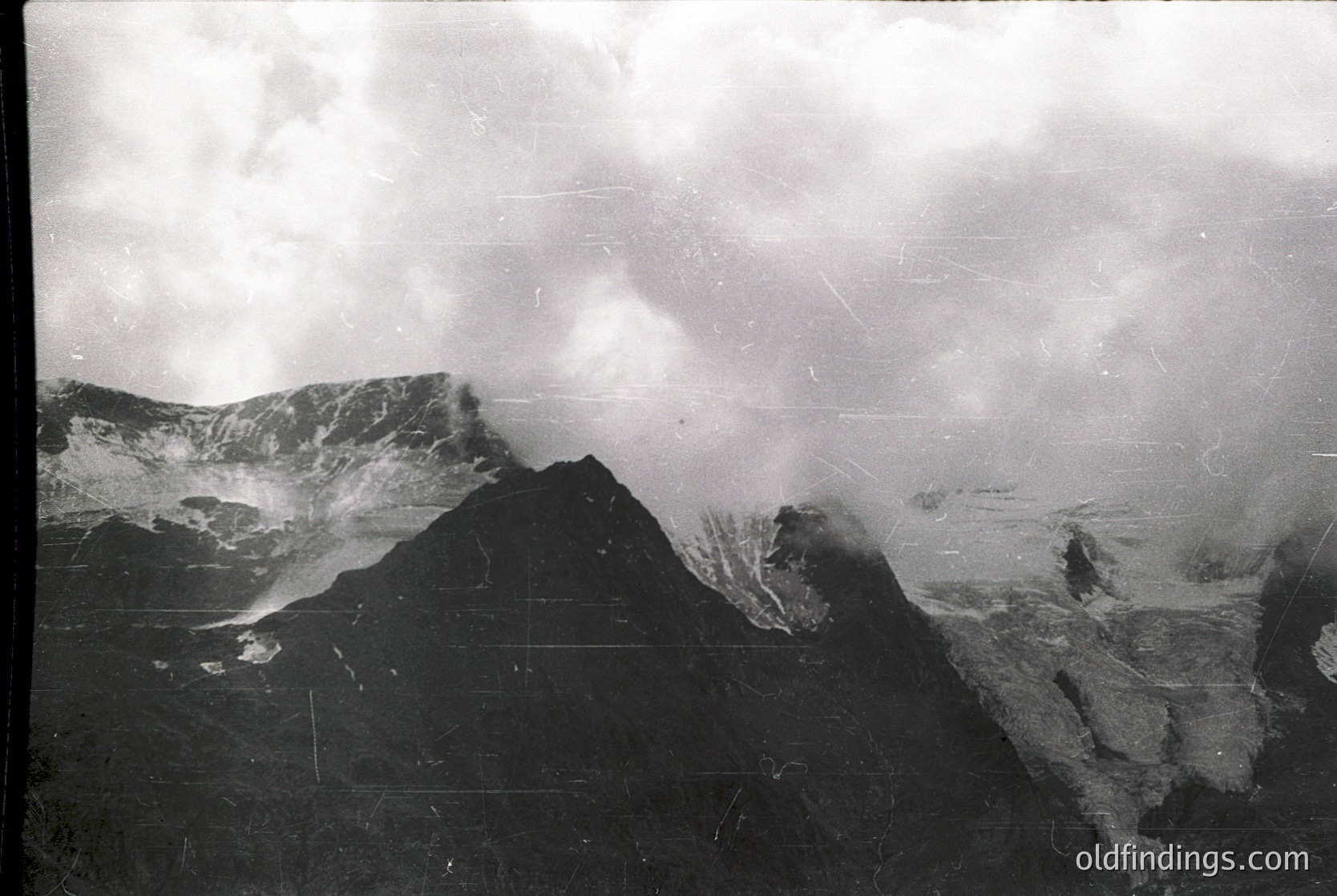 Vintage black-and-white aerial shot of rugged alpine terrain with steep peaks and mist-covered slopes. Likely mid-20th century due to grain and contrast.