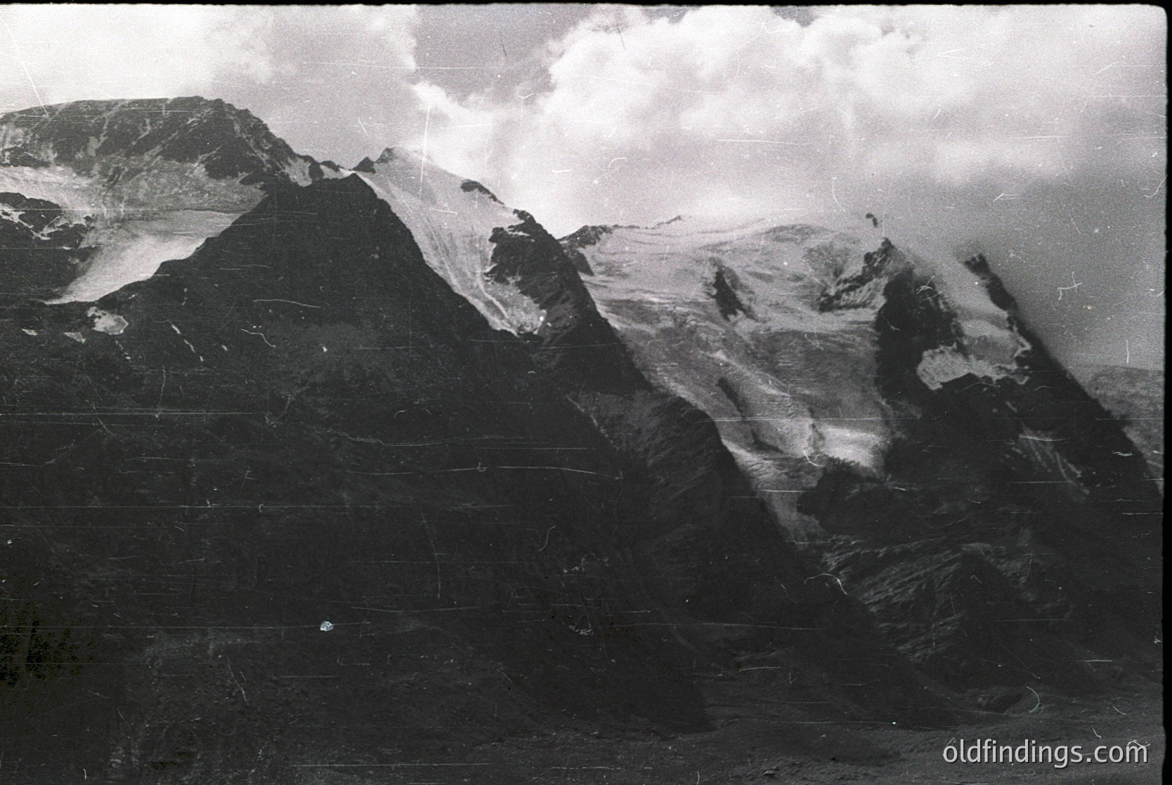 High-contrast black-and-white alpine landscape featuring jagged peaks and snowfields. Dramatic cloud cover obscures mid-mountain ridges, emphasizing verticality and rugged terrain. Likely mid-20th century due to grain and tonality.