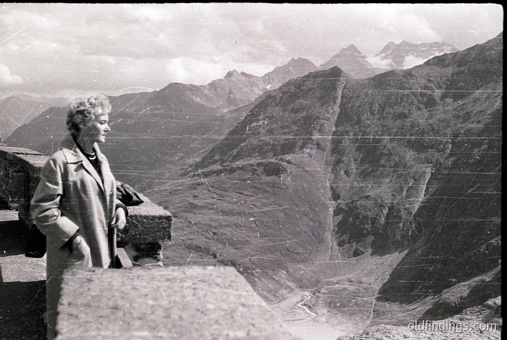 Mid-century woman in a structured coat poses atop rugged alpine ridge, framed by jagged peaks. Vintage monochrome print suggests or European Alps. Dramatic lighting highlights rocky terrain and vertical cliffs.