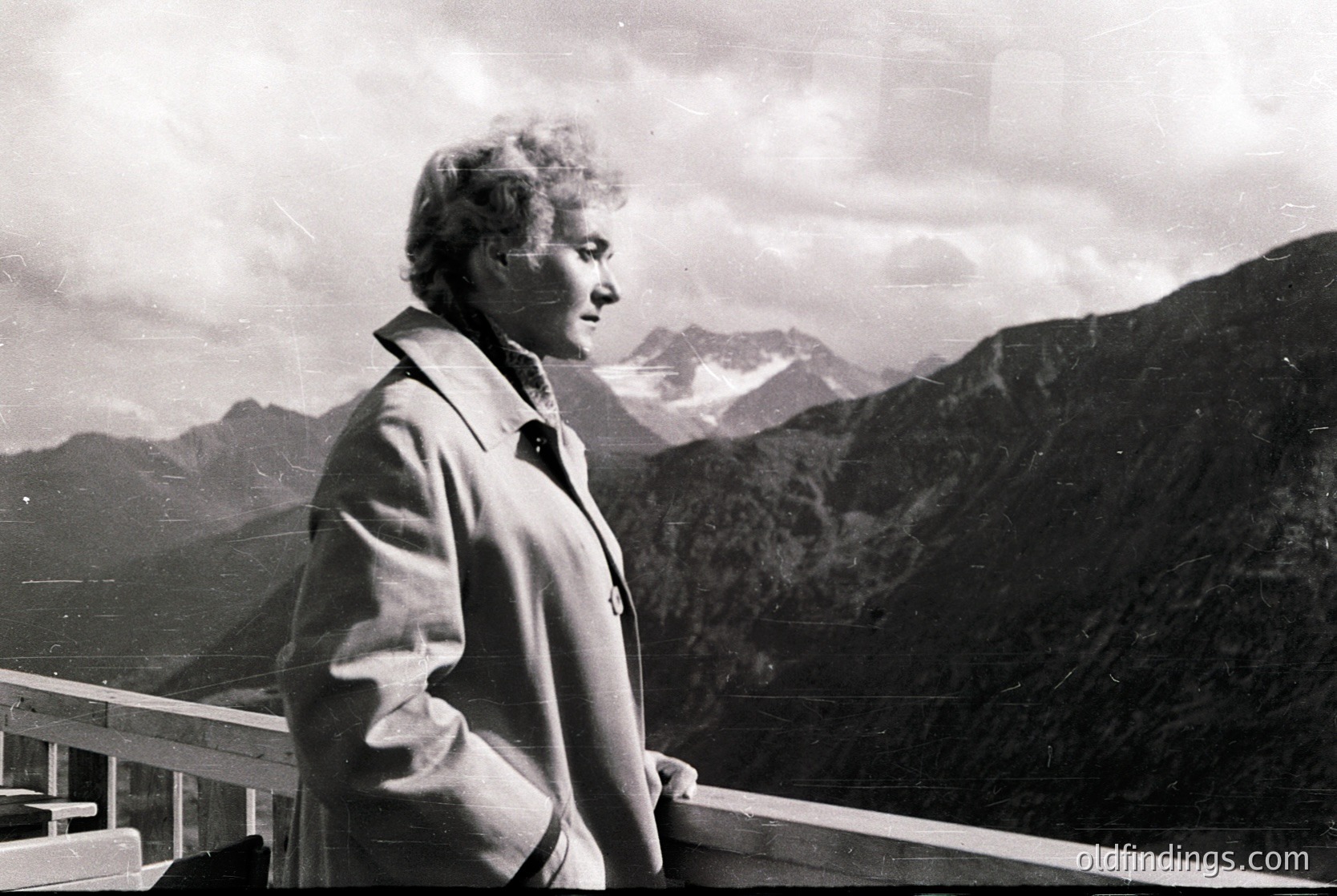 Mid-century woman in a collared coat stands on a metal railing overlooking rugged alpine peaks, suggesting mid-20th century outdoor travel. Dramatic cloud cover enhances the mountainous backdrop.