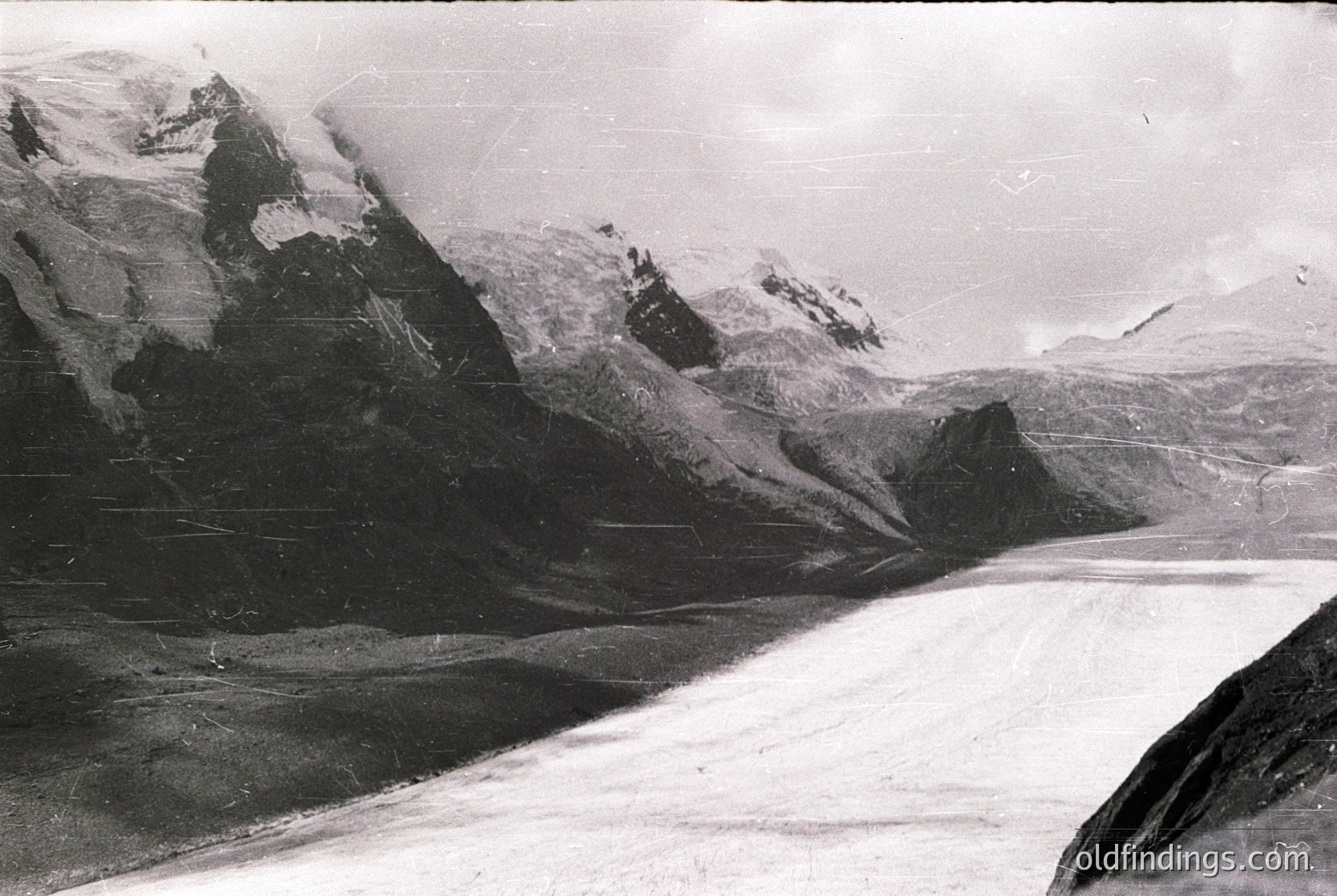 Vintage black-and-white alpine glacier scene with jagged peaks and snow-covered slopes. Evidence of early 20th-century photography via grain and sepia tone. Dramatic lighting highlights ice formations and rugged terrain.
