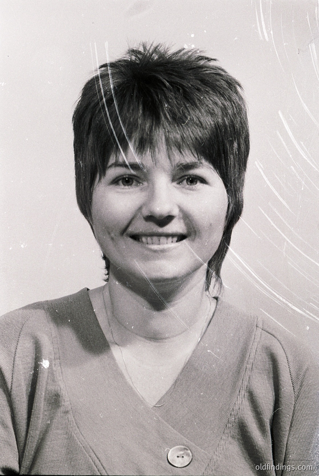 Portrait of a woman with short, textured hair and a warm smile, wearing a V-neck sweater. Style suggests mid-20th century (1950s–1970s). Blurred background indicates studio lighting.