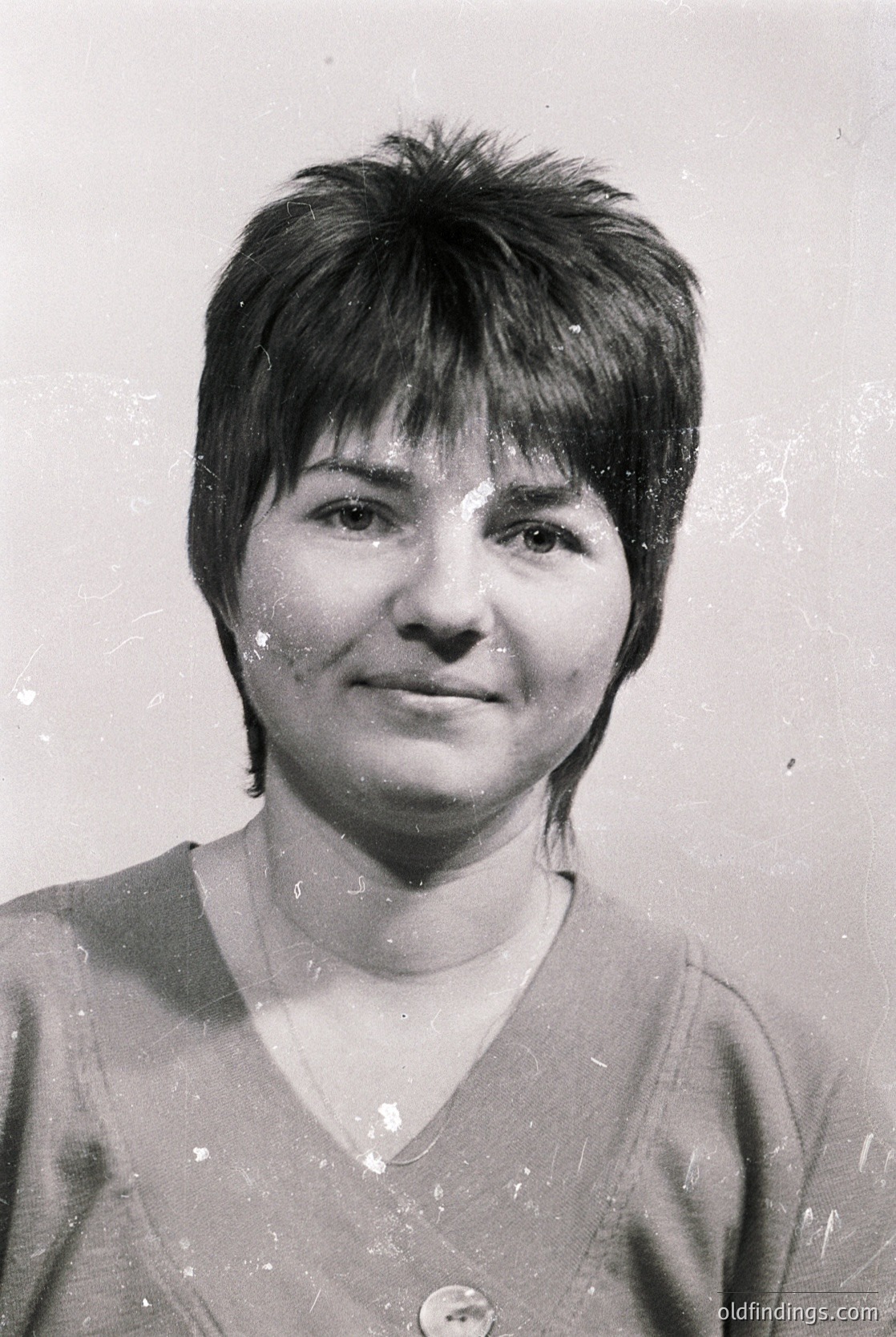 Portrait of a woman with short, angled bangs and a neutral expression, likely taken in the 1970s–1980s. She wears a simple V-neck sweater and a small, round brooch at the collar. The grainy, sepia-toned background suggests a studio or formal setting.