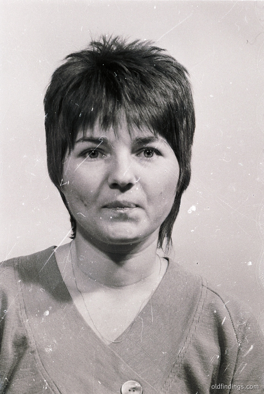 Portrait of a woman with a short, angled bob hairstyle, taken in black-and-white. She wears a simple V-neck sweater and a small, round pin on her lapel. The image appears aged with visible scratches and fading, suggesting it’s a vintage or archival photo.