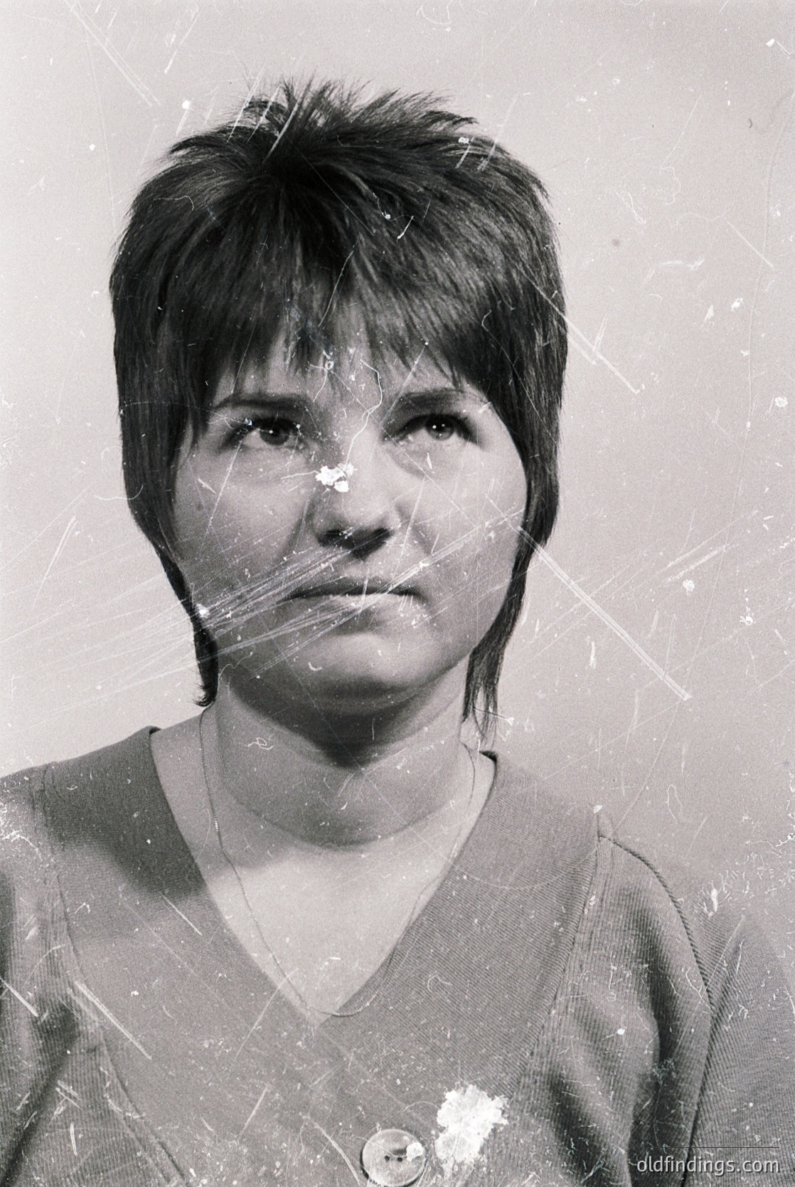 Portrait of a woman with short, straight hair and a neutral expression, captured in black-and-white. She wears a simple V-neck sweater and a small, round badge on her left lapel. The image shows visible scratches and damage, suggesting it may be an old or scanned document.