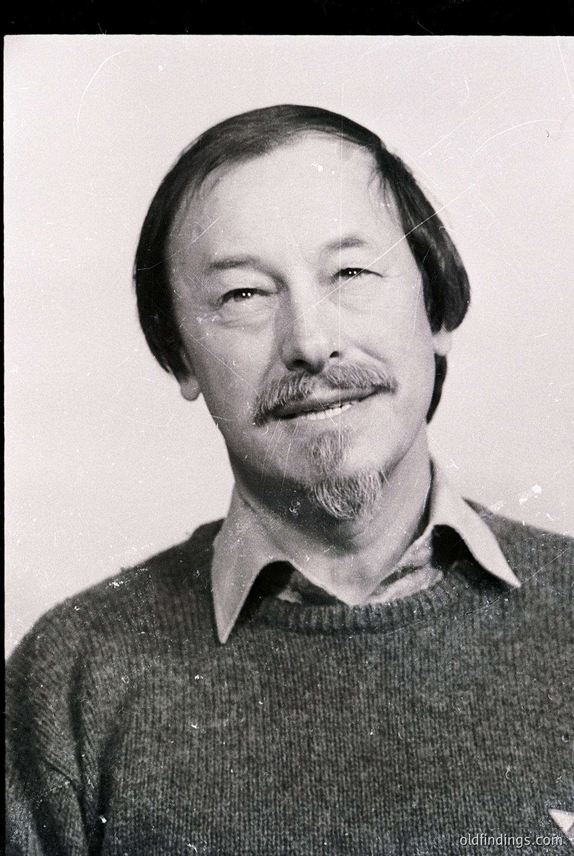 Black-and-white portrait of a man with a mustache and beard, wearing a collared sweater. Mid-century styling suggests 1960s–1970s. Neutral expression, plain background.