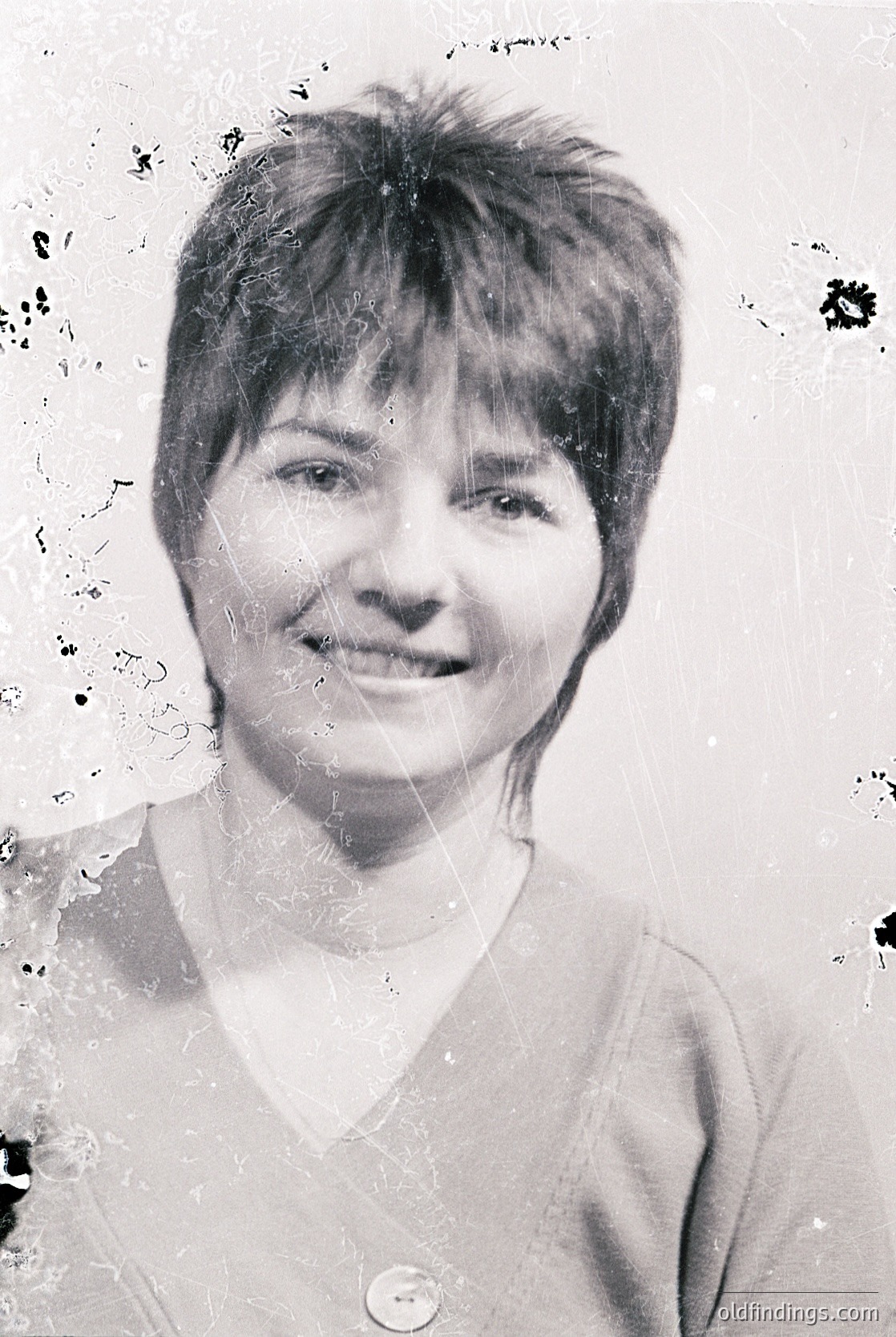 Vintage black-and-white portrait of a woman with short, voluminous hair and a warm smile. She wears a simple V-neck top with a small, round button. The image shows signs of aging (tears, stains) and a soft focus effect, suggesting mid-20th century photography.