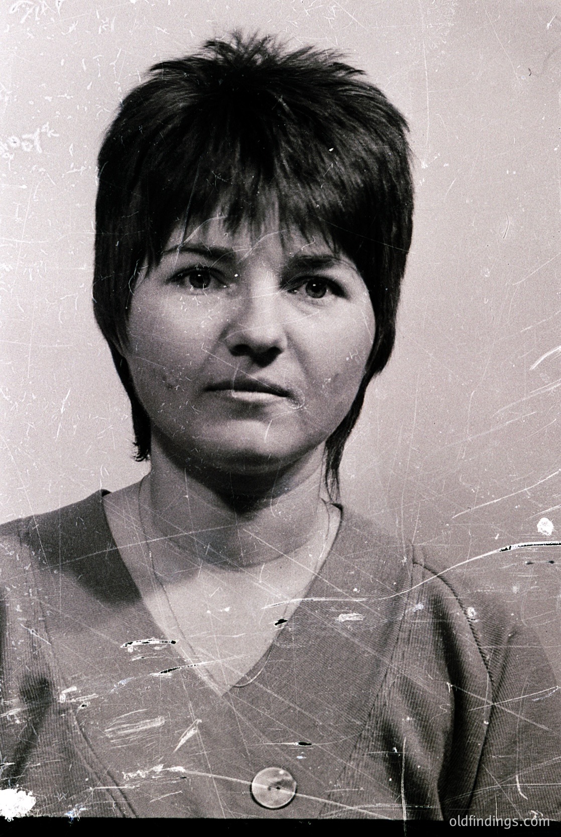 Vintage black-and-white portrait of a woman with short, straight hair and a neutral expression. She wears a simple V-neck top with a visible button. The image shows signs of aging—cracks and wear on the edges—suggesting it may be a scanned or printed document from the mid-20th century.