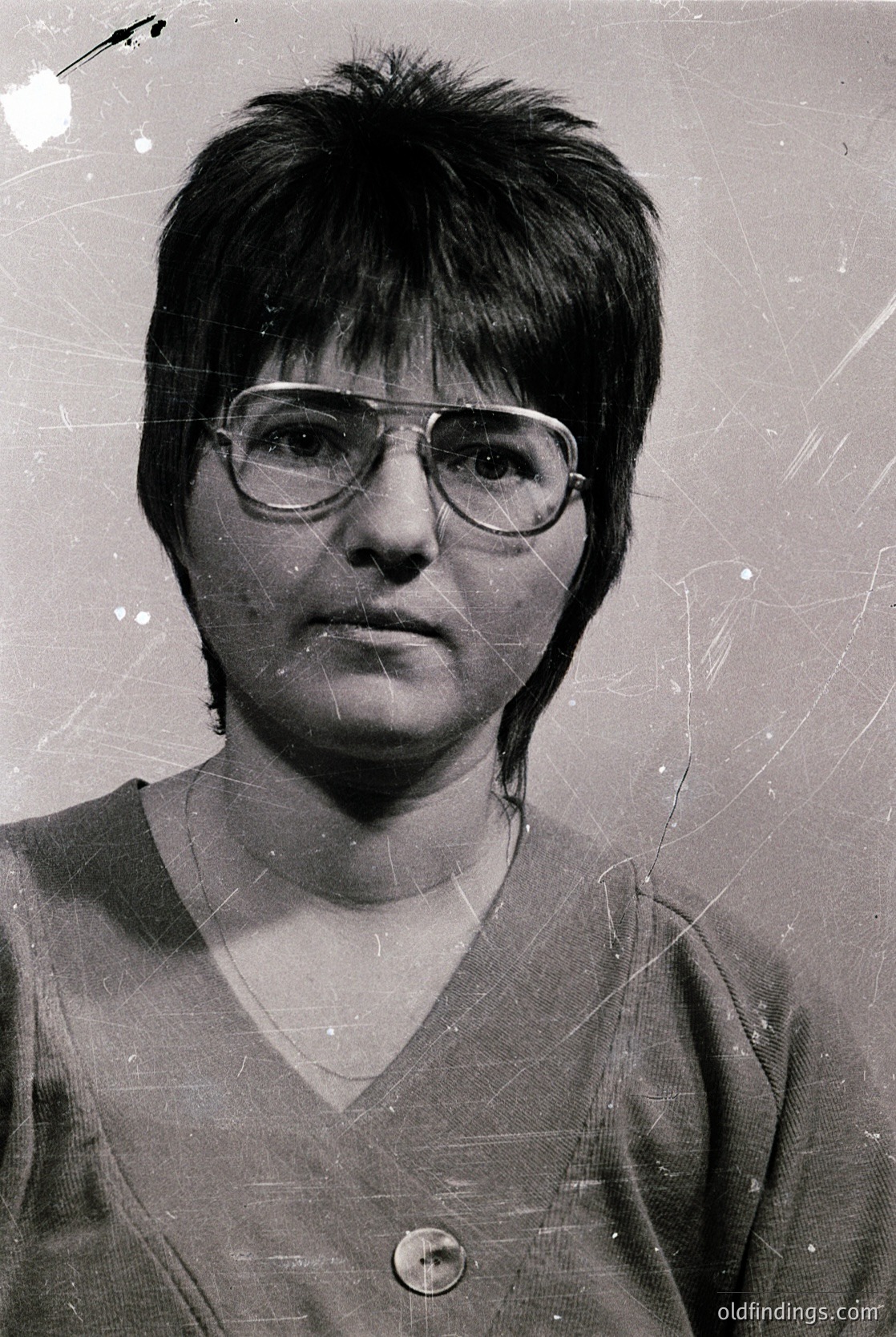 Black-and-white portrait of an individual with short, textured hair and rectangular glasses, wearing a buttoned V-neck sweater. The aged, slightly scratched film suggests a mid-20th century origin ( ). Neutral expression and formal pose indicate possible identification or documentation use.
