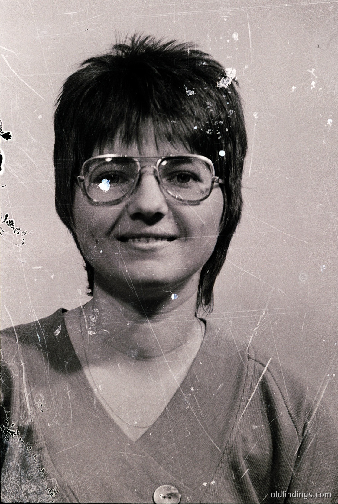 Vintage portrait of a person with short, textured hair and round glasses, wearing a V-neck sweater. The image shows signs of age with visible scratches and creases. Likely from the mid-20th century (1950s–1970s).