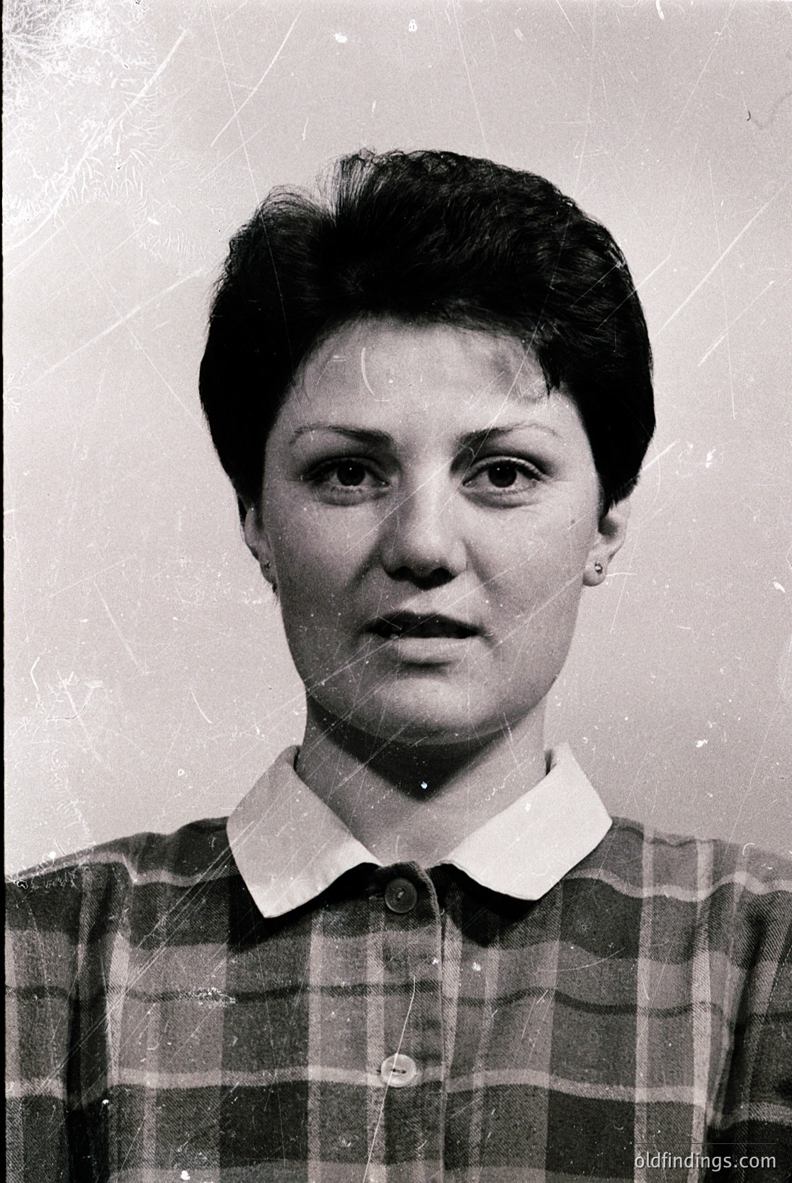 Portrait of a woman in a mid-century plaid blouse, short hairstyle, and minimal jewelry—likely a 1960s–1970s identification or passport photo. Neutral expression, plain background suggests institutional use.