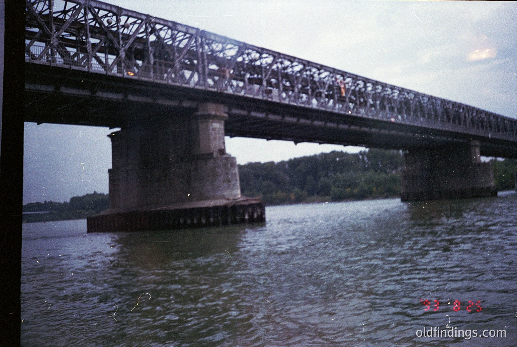 Vintage steel truss bridge spanning a river, supported by concrete piers. Crowds of people walking across, suggesting mid-20th century urban transport. Overcast sky and blurred greenery in background. Likely or industrial-era infrastructure.