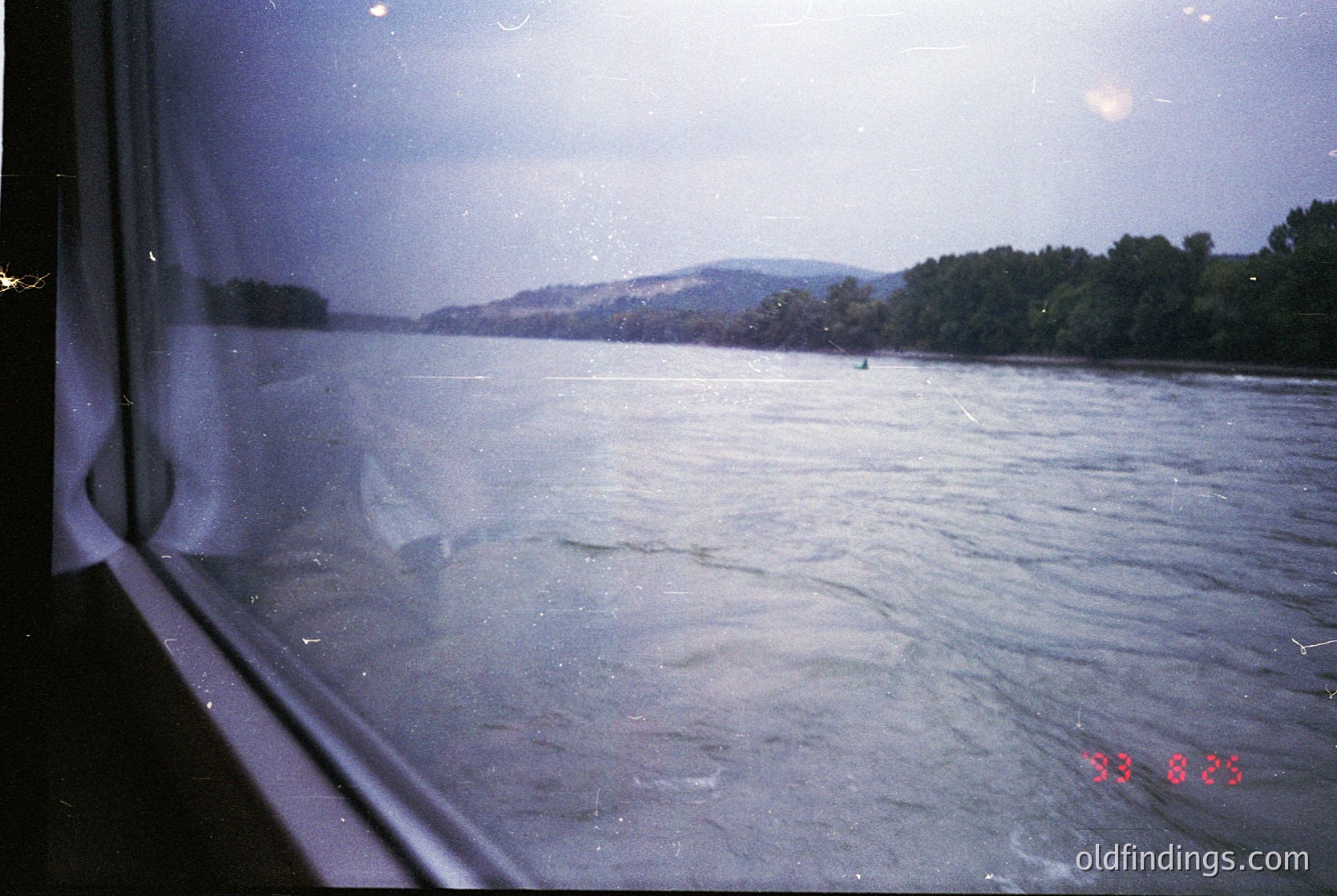 Vintage view through a train window showing turbulent river waters, likely the Danube, with distant forested hills. Overcast sky and blurred motion suggest mid-20th century travel.