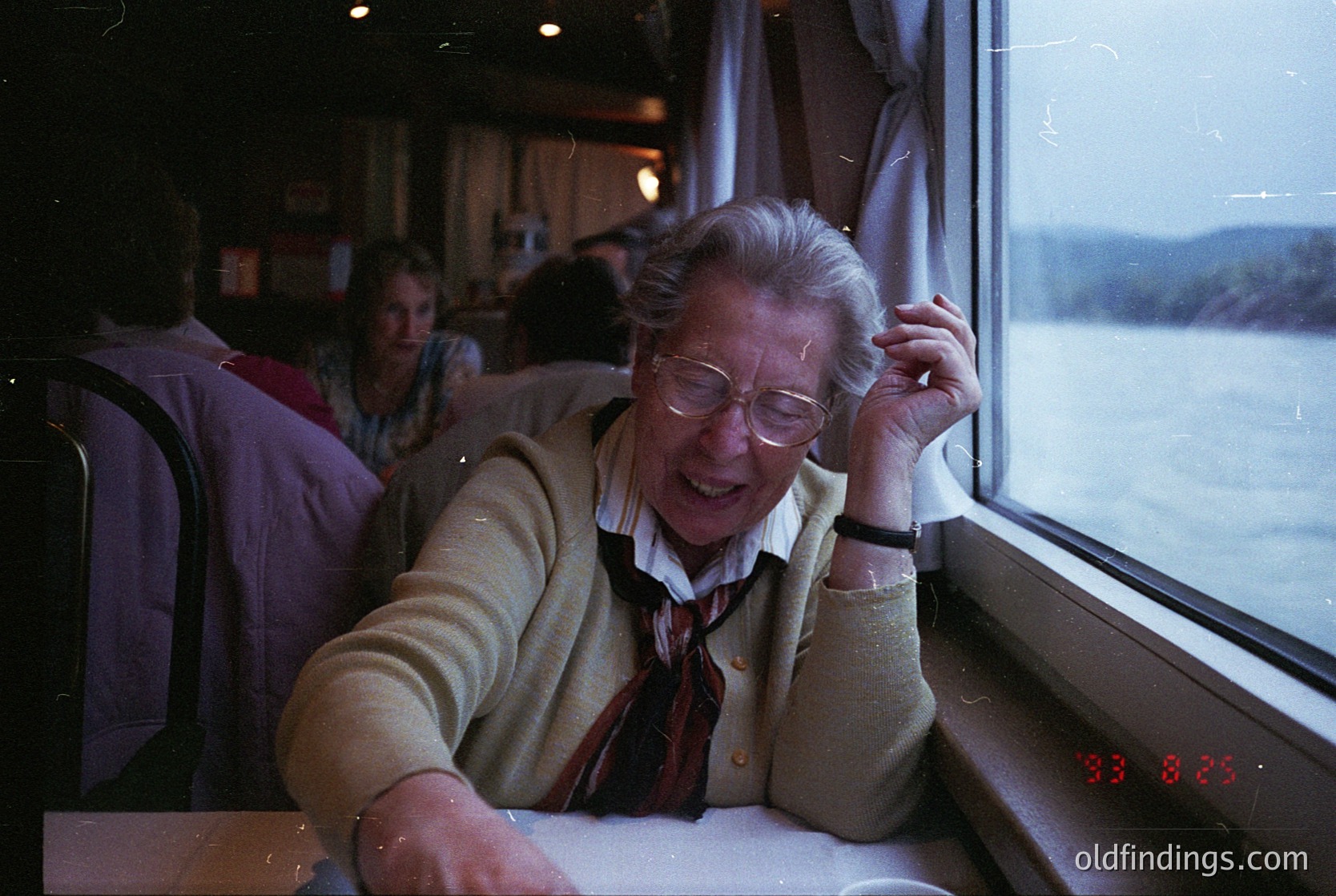 Vintage train interior shot featuring an elderly woman in a beige cardigan and red scarf, leaning on a window sill with a relaxed smile. Outside, a scenic lake and forested hills are visible. Digital clock displays "33:88:33". Mid-20th century travel aesthetic.