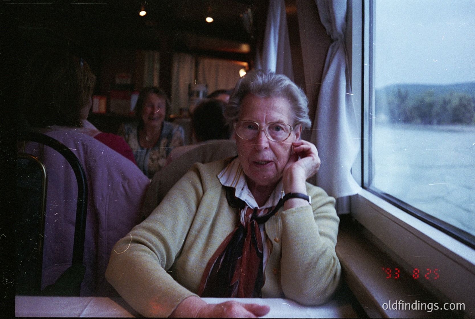 Vintage train interior shot featuring an elderly woman in a cream sweater and red scarf speaking on a corded phone, seated near a window overlooking a lake. Wood-paneled walls and blurred passengers in the background suggest mid-20th century travel.