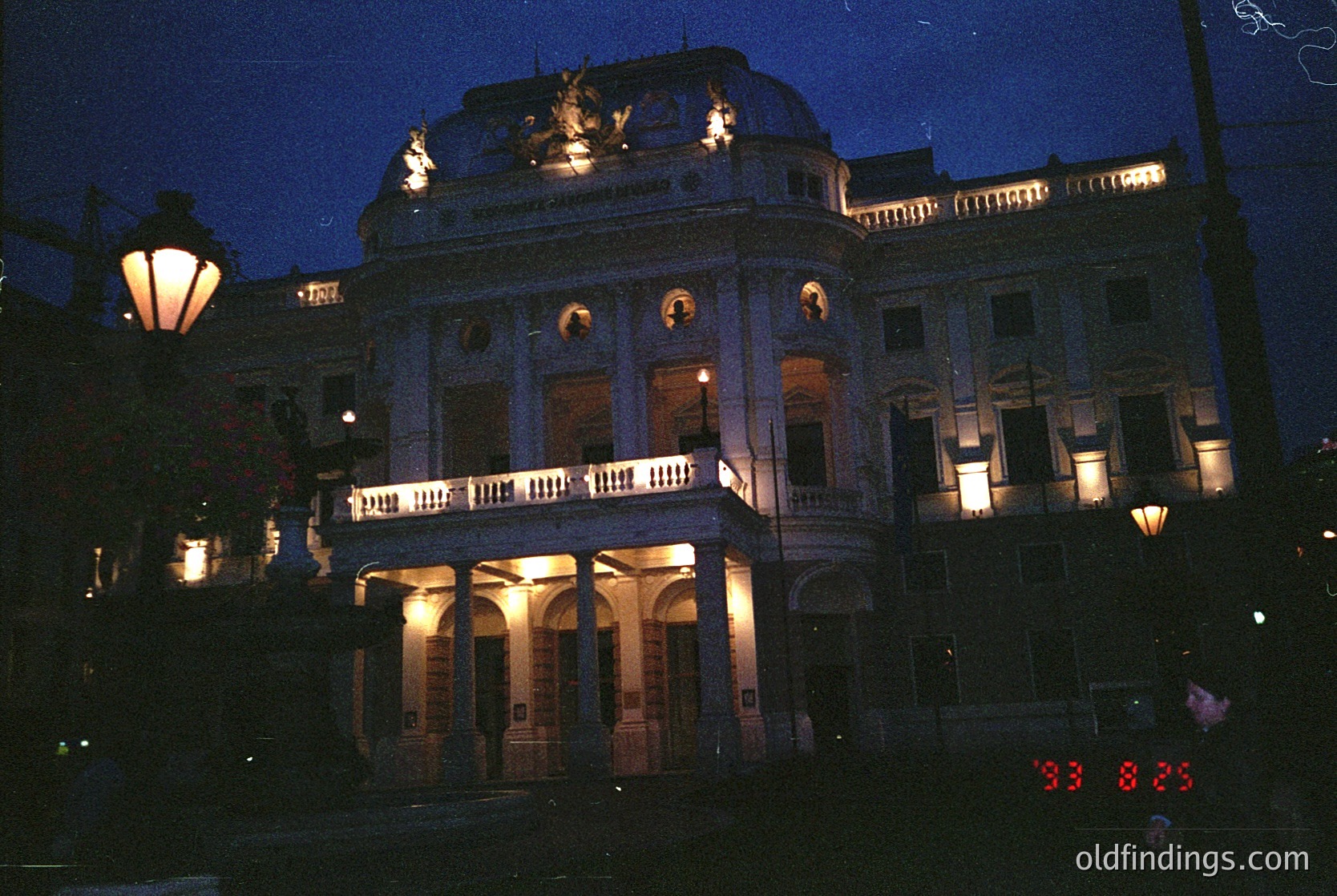 Neoclassical building illuminated at dusk, featuring arched windows, balustrades, and ornate rooftop details. Warm lighting highlights architectural symmetry. Likely European, late 19th to early 20th century.