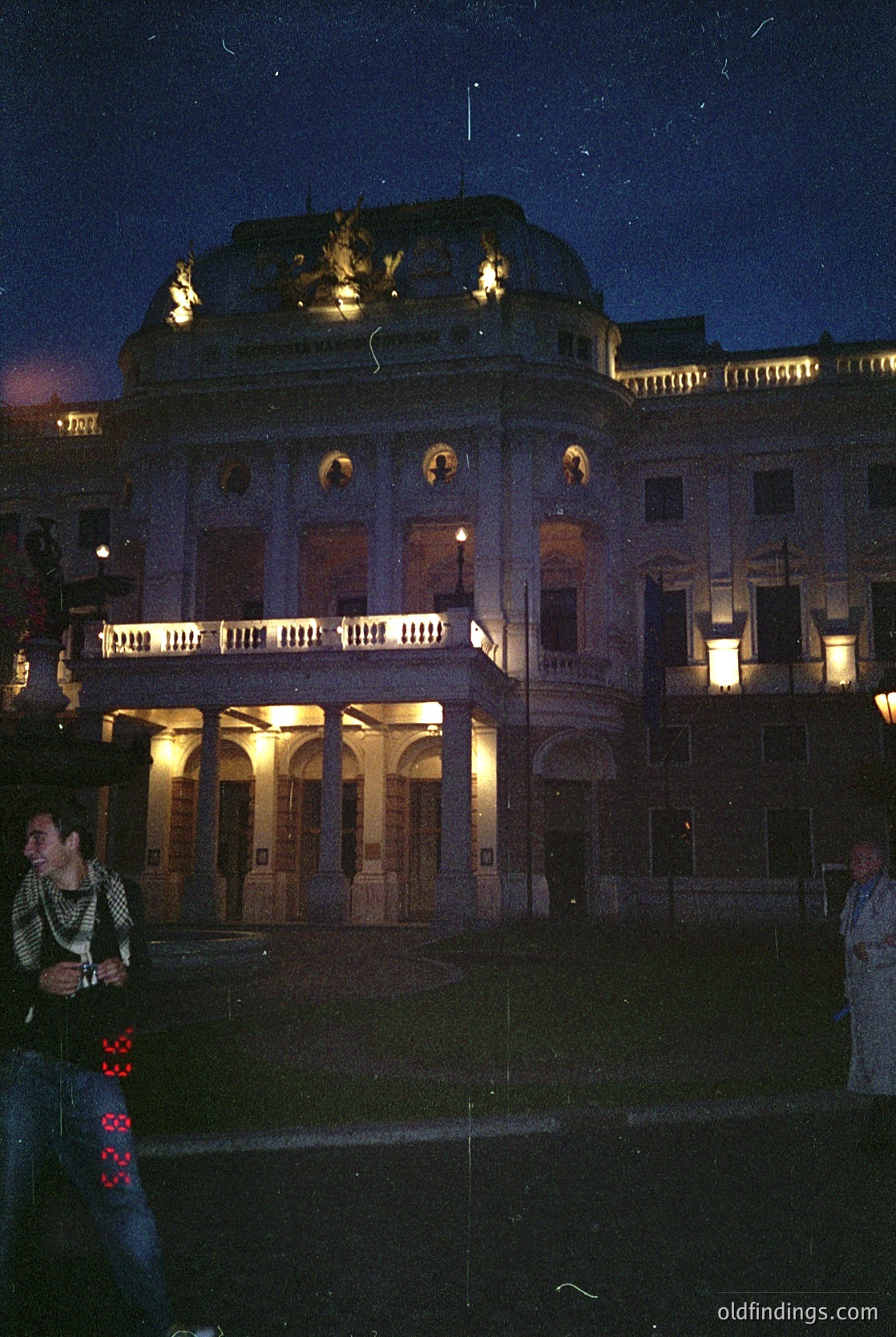 Neoclassical building illuminated at dusk, featuring symmetrical arches, columns, and a central pediment with sculptural relief. Warm lighting highlights architectural details like balustrades and decorative cornices. Two silhouetted figures stand in the foreground, one holding a camera. Likely a public or cultural landmark.