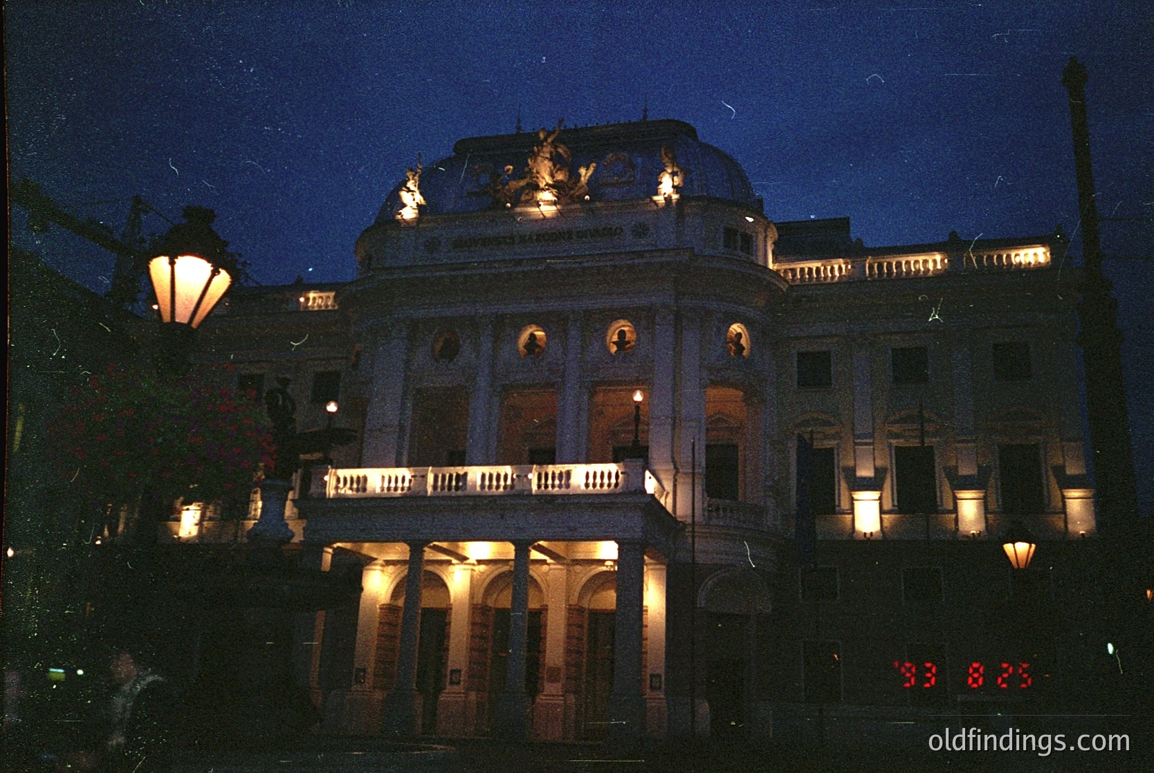 Neoclassical building illuminated at dusk, featuring symmetrical columns, arched windows, and decorative balustrades. The central section includes a prominent pediment with sculptural relief. Warm street lamps enhance architectural details. Likely a cultural or governmental structure from the 20th century.