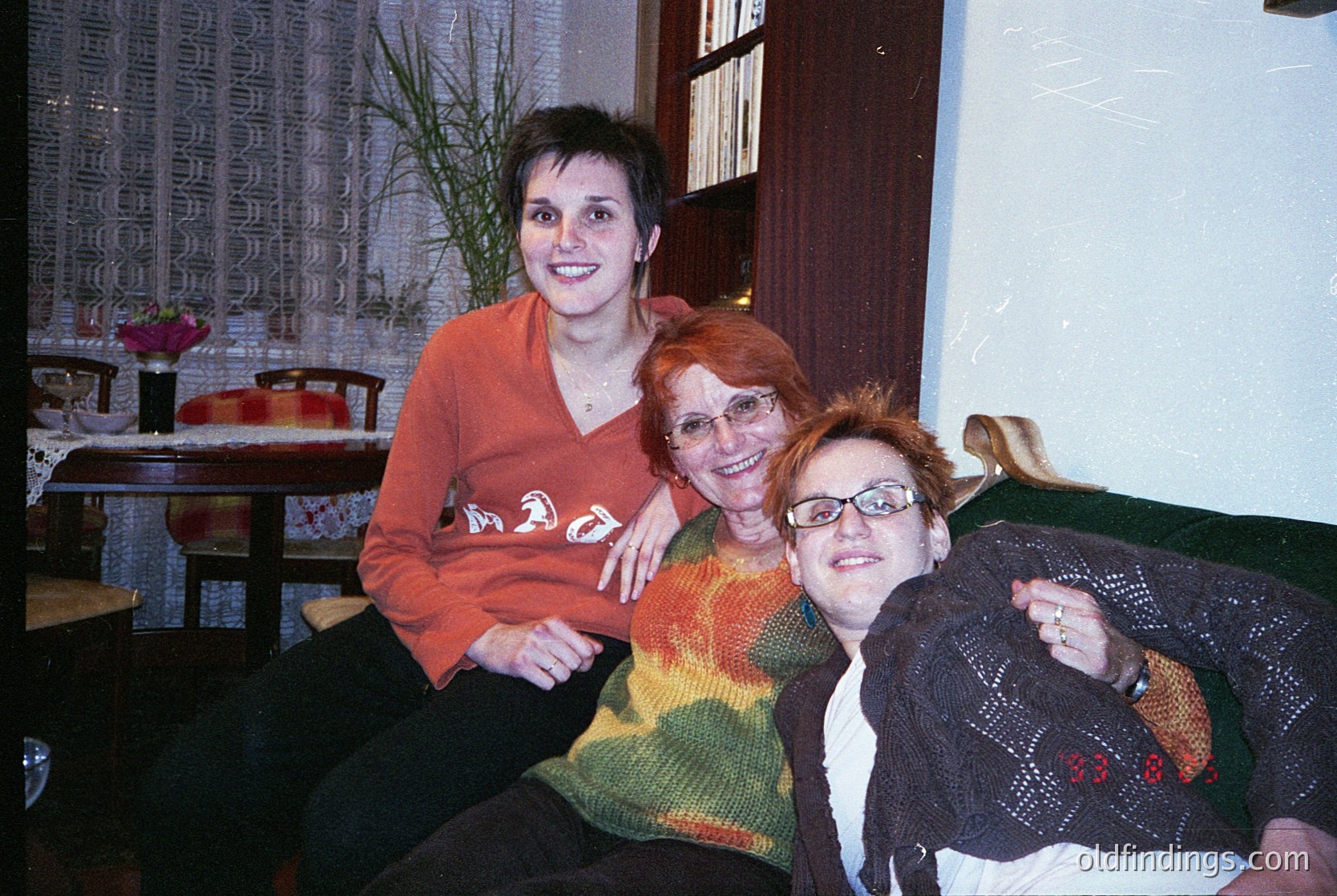 Vintage indoor portrait of three women in a cozy, retro-style café. The woman on left wears an orange sweater with "MAD" printed in bold letters; center wears a multicolored knit sweater; right holds a blanket with "33 8" embroidered. Wooden tables, floral curtains, and a potted plant add warmth. Likely late 20th century, possibly Eastern European.