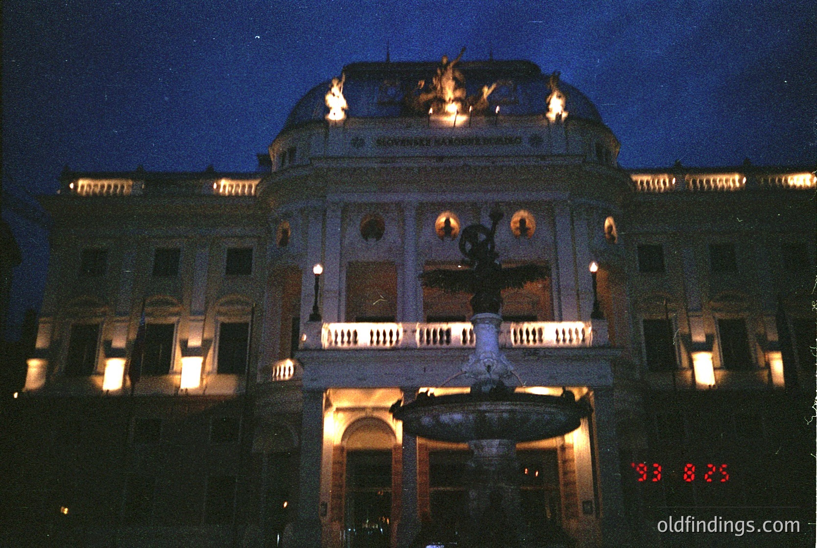 Neoclassical building illuminated at dusk, featuring ornate columns, balustrades, and a central fountain. The inscription "Slavianski Bazar" suggests Bulgarian cultural heritage, likely Sofia’s iconic Slavianski Bazar Hotel. Architectural details include symmetrical windows, decorative urns, and warm lighting highlighting its grandeur.