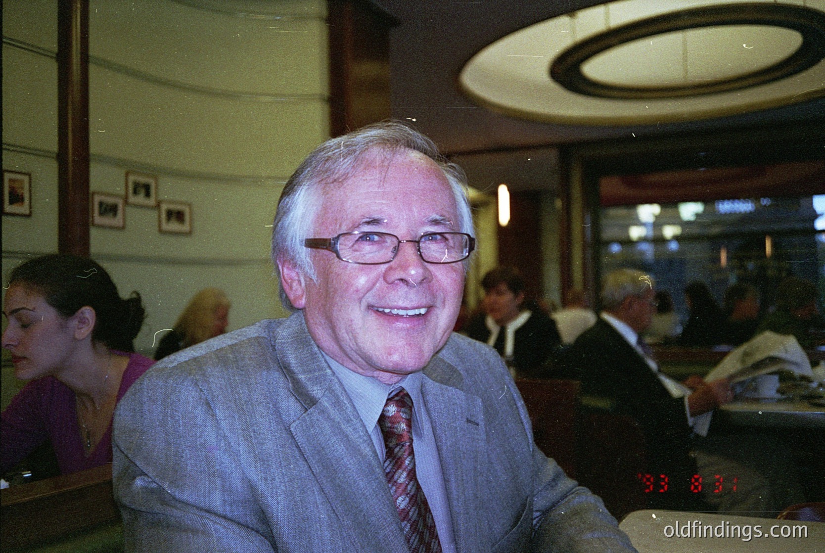Mid-century café portrait: Man in 1970s-style suit, glasses, and striped tie, seated indoors with curved ceiling and framed art. Warm lighting and blurred patrons suggest a lively social setting. éCulture