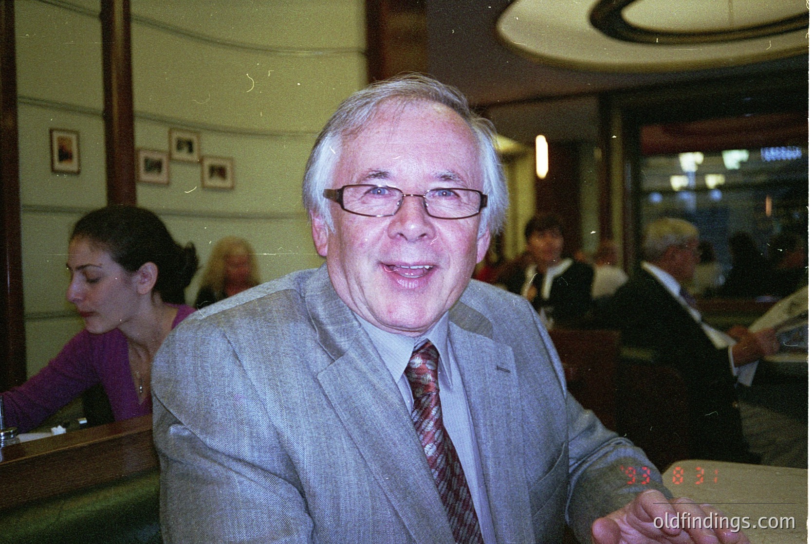 Classic 1990s-era indoor portrait in a formal setting. Man in a light grey suit, red-striped tie, and glasses poses with a playful expression. Wood-paneled walls, framed artworks, and blurred attendees suggest a conference or business event. Digital camera timestamp reads "9:36 33".