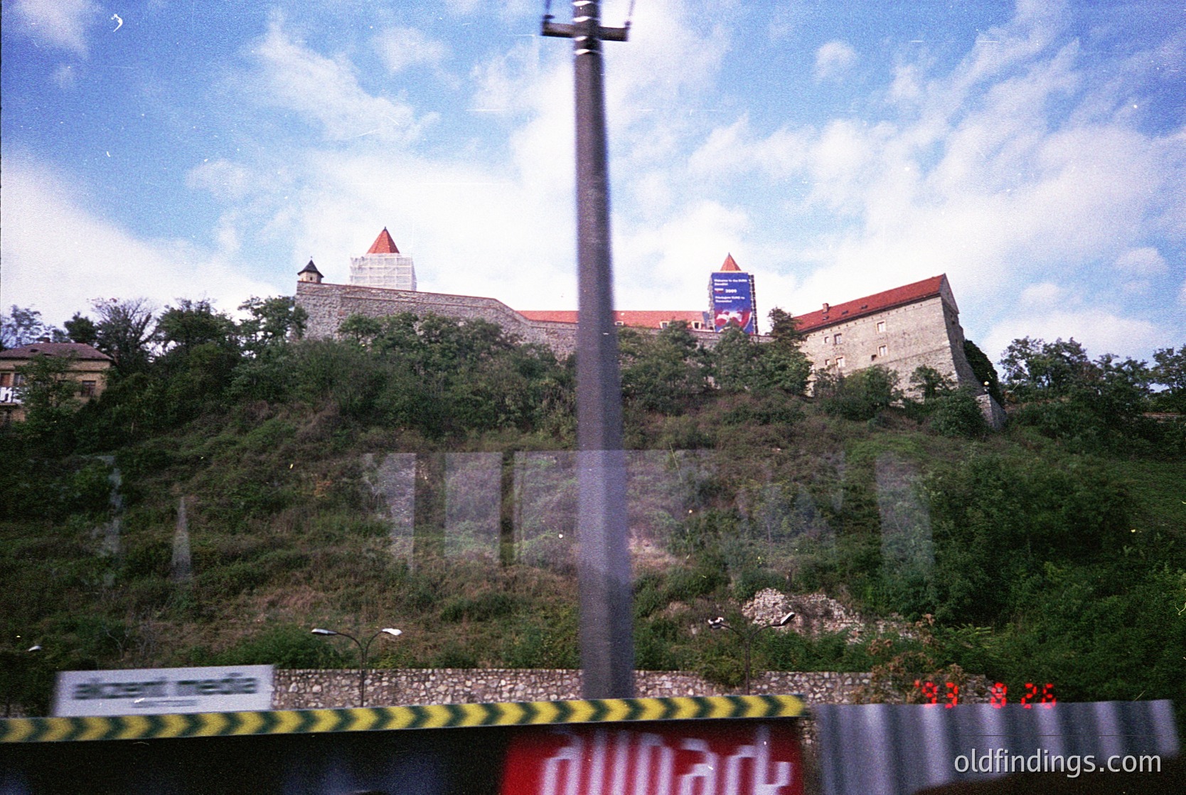 Historic hilltop fortress with medieval architecture, featuring stone walls and conical towers. Overgrown vegetation surrounds the base. Image taken from a bus window, indicated by blurred motion and timestamp "19:33:25" on the metal barrier. Likely Eastern European setting.