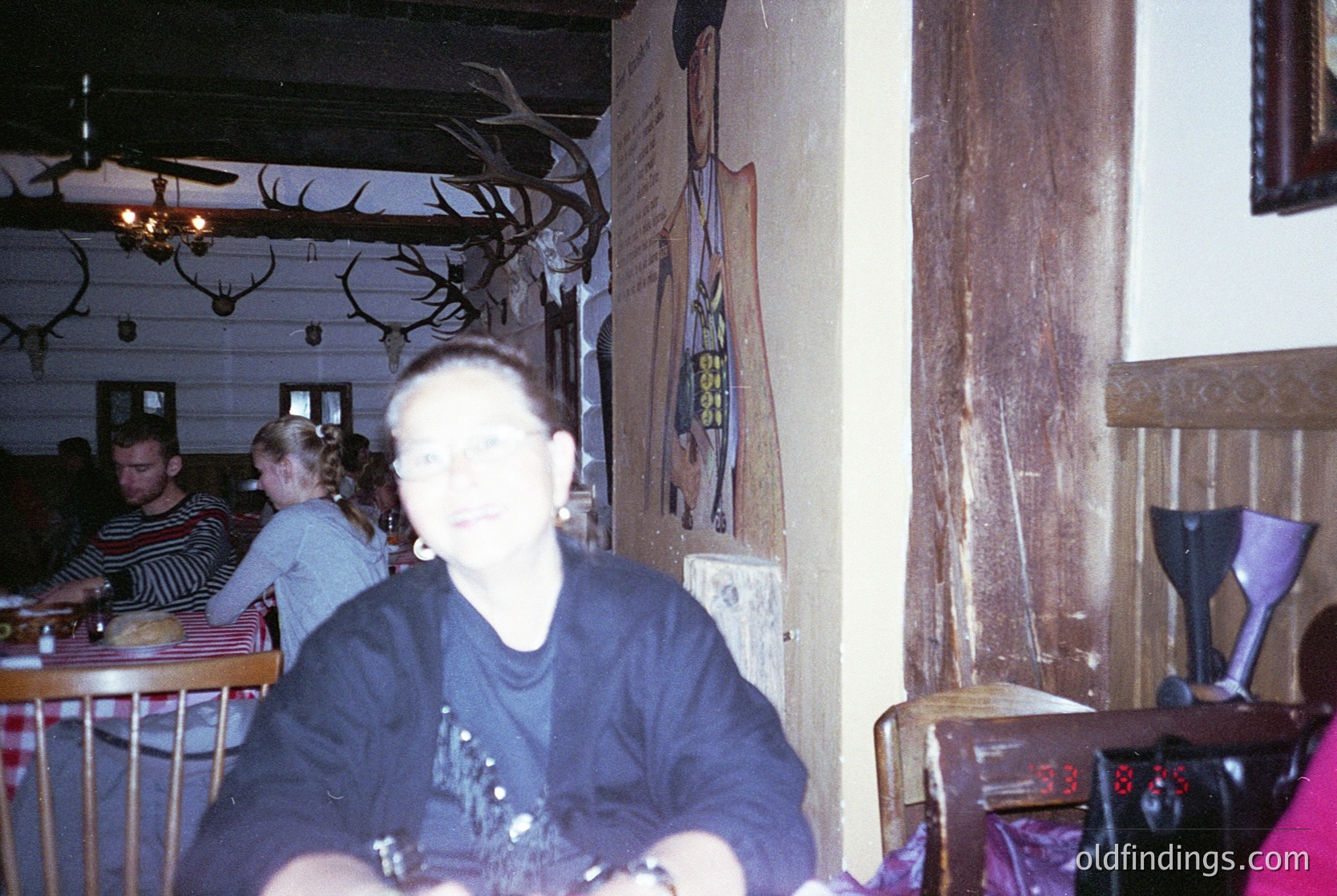 Vintage indoor dining scene with rustic wooden decor. A central figure wears a dark long-sleeve shirt with a graphic print, seated at a wooden table with a blurred menu. Deer antlers and framed posters adorn the walls, suggesting a lodge or mountain-themed café. Other patrons in casual 1990s attire dine in the background. Warm lighting and a chandelier enhance the cozy ambiance. é
