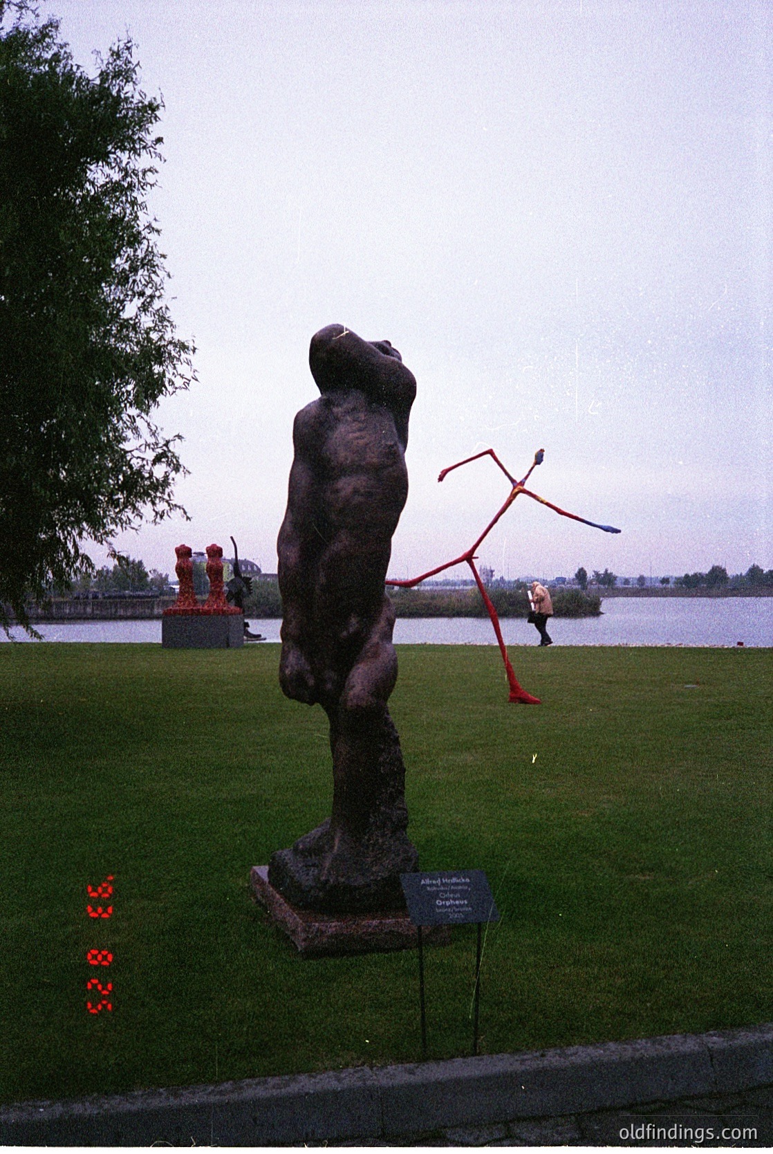 Bronze abstract sculpture of a seated figure with outstretched arm in a landscaped park, near a body of water. Red kinetic sculpture in background. Plaque identifies artist and title ("Organism"). Overcast sky and greenery suggest a public outdoor space.