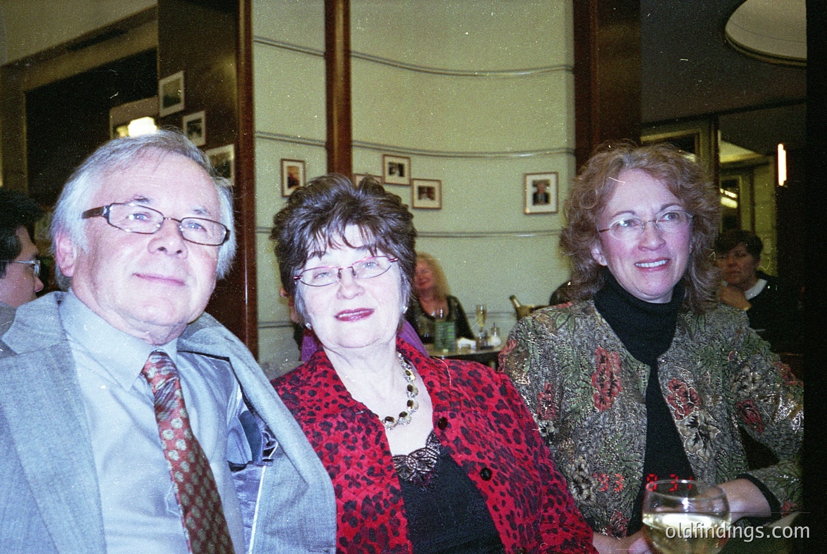 Three adults pose indoors in a formal setting, likely a 21st-century event. The man wears a patterned tie and glasses; the woman in the center sports a leopard-print blouse and necklace; the woman on the right holds a wineglass and wears a patterned jacket. Wood-paneled walls and framed art suggest a classic venue. [Indoor group portrait at a formal event, featuring patterned attire and vintage decor ]