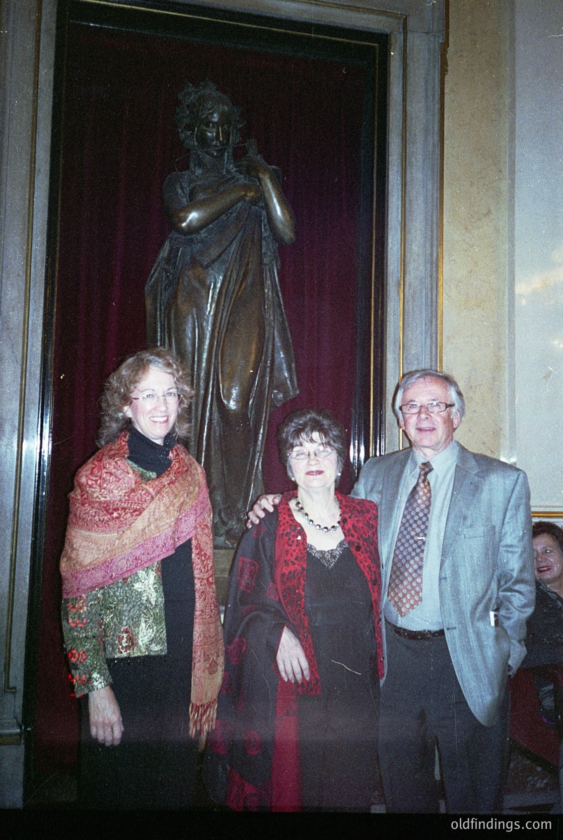 Three individuals pose indoors beside a classical bronze statue of a draped figure in a grand hall, likely a museum or cultural institution. The woman on the left wears a patterned shawl over a floral blouse; the central figure in a red dress with pearl jewelry; the man on the right in a suit with a checkered tie. Formal attire suggests an event from the **1970s–1980s**.