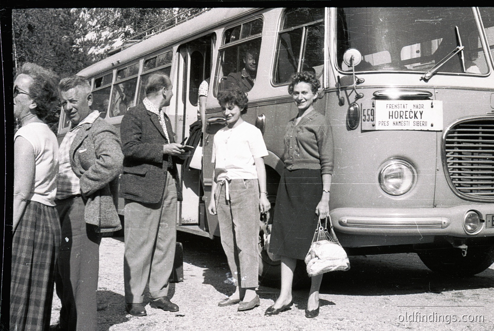 Mid-20th century group boarding a vintage Czech bus, marked "Horecky" with route details. Men in suits, women in 1950s-60s attire (pencil skirts, cardigans). Outdoor setting with greenery and gravel road.