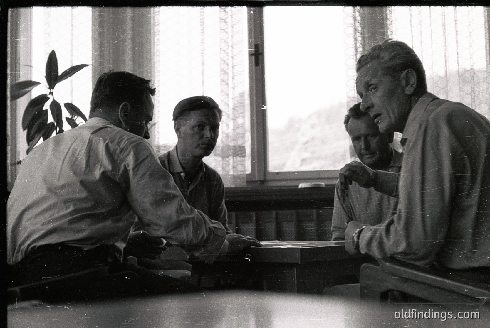 Four men seated around a small wooden table in an indoor setting, likely a café or lounge, playing cards. Mid-20th century attire suggests or . Natural light filters through curtains, revealing a blurred outdoor scene. Indoor social gathering with vintage ambiance.