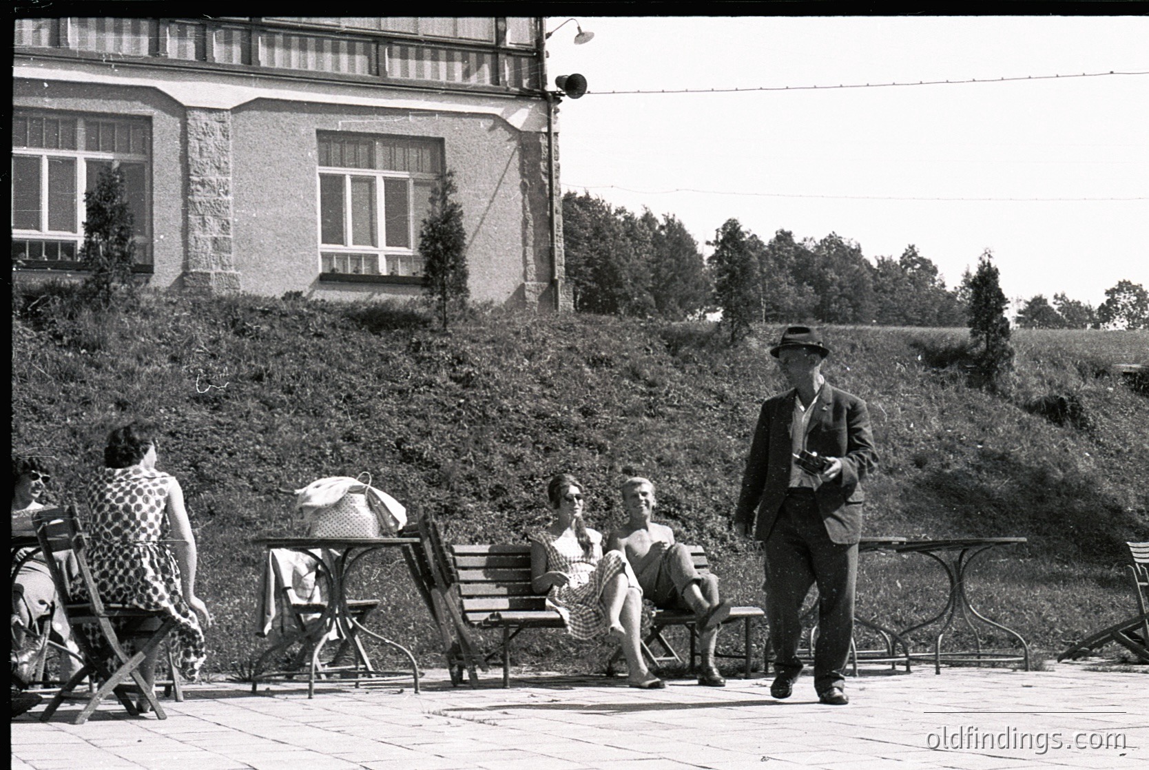 Mid-20th century urban park scene featuring vintage metal benches, stone building with large windows, and lush greenery. A man in a fedora and suit stands holding a rolled document, while others sit casually in checkered and striped clothing. Black-and-white, suggesting 1950s–1960s era.
