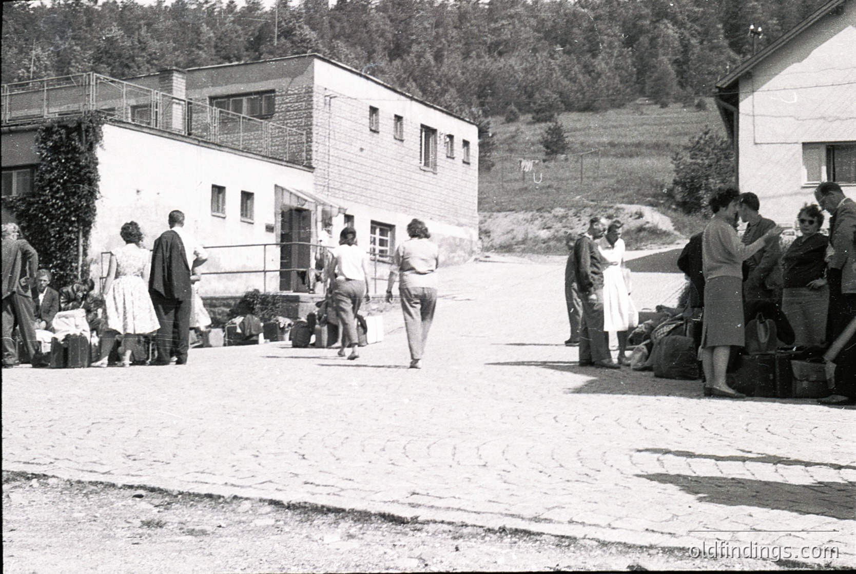 Mid-20th century rural gathering near modernist brick buildings with flat roofs and balconies. Group of people—men in suits, women in dresses—standing, walking, and conversing outdoors. Luggage and bags suggest travel or a communal event. Lush greenery and forested hills in background. Likely European countryside, 1950s–1960s.