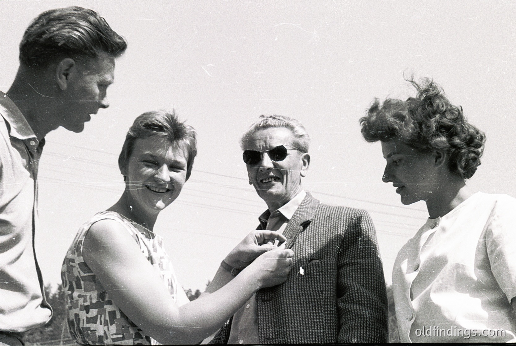 Four individuals pose outdoors in 1960s-era attire—men in suits, women in patterned dresses and hairstyles. The man in sunglasses adjusts a woman’s blouse, suggesting a social or celebratory occasion. Bright daylight and blurred background indicate an informal, candid moment. Potential historical context: Western Europe or North America. [Mid-century social gathering with 1960s fashion and candid poses ]