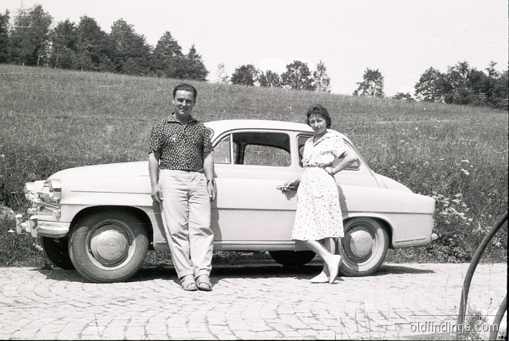 Mid-century classic car (likely a Fiat 1100/1200 series) with vintage white paint and wire-spoke wheels, parked on a cobblestone driveway. Two individuals pose beside it—man in patterned shirt and rolled trousers, woman in floral dress and sandals. Rural countryside background with open fields and trees, suggesting a European setting. Likely 1950s–1960s.