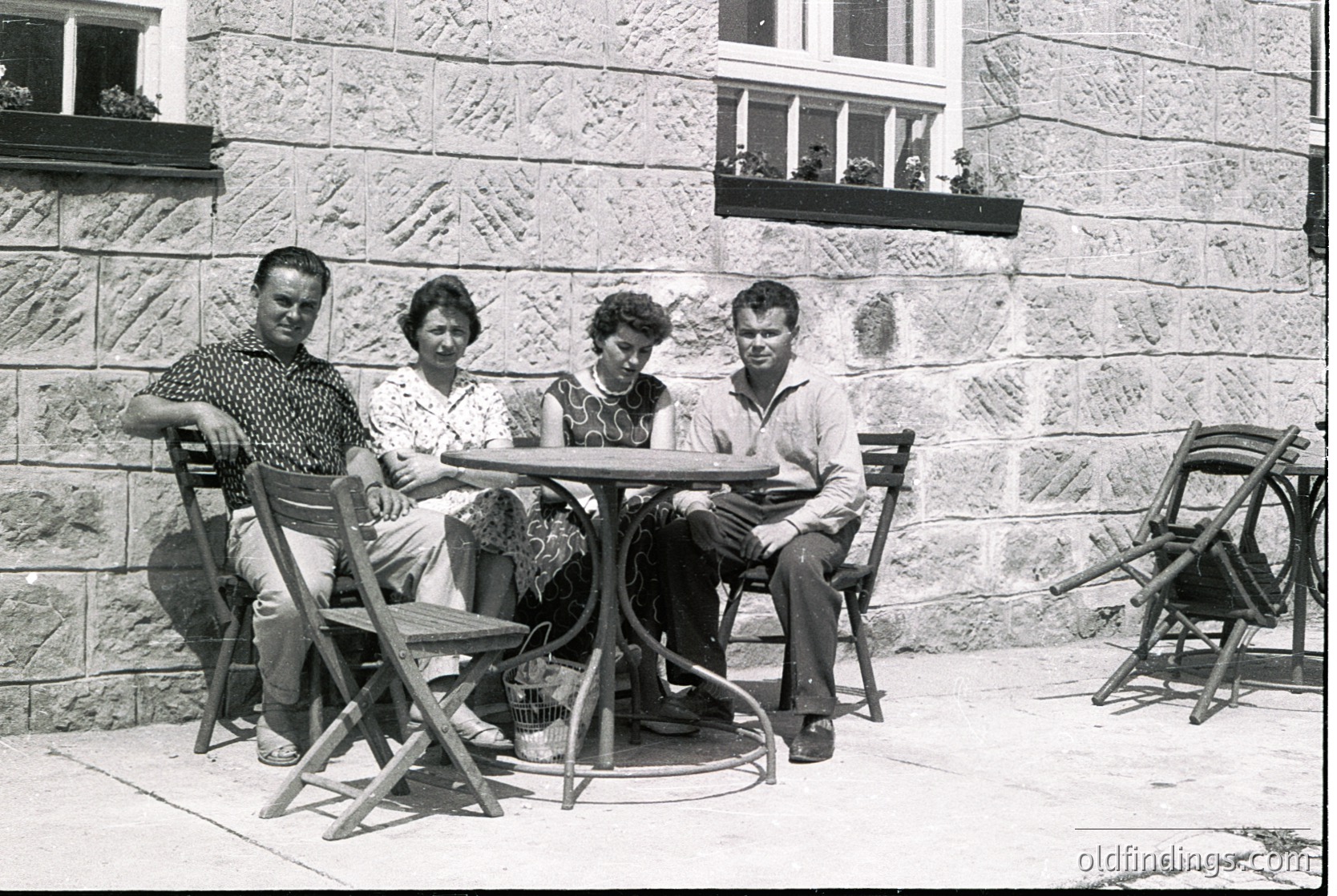 Four individuals pose outdoors at a round metal table, seated on vintage folding chairs against a stone wall with flower boxes. Mid-century clothing (men in button-downs, women in patterned dresses) suggests a 1950s–1960s European setting. The composition captures a relaxed, informal gathering.