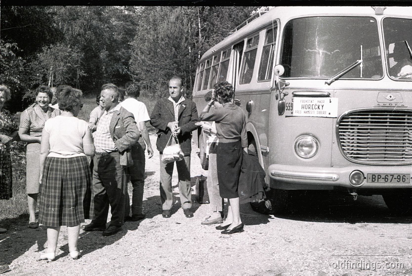 Vintage group boarding a **Horecky** branded bus (license plate BP-67-96) in mid-20th century, likely 1960s. Six adults in period clothing—plaid skirts, cardigans, and suits—pose near a forested roadside. Bus features classic rounded design with "Sovak" branding. Rural European setting, possibly Czechoslovakia or Slovakia.