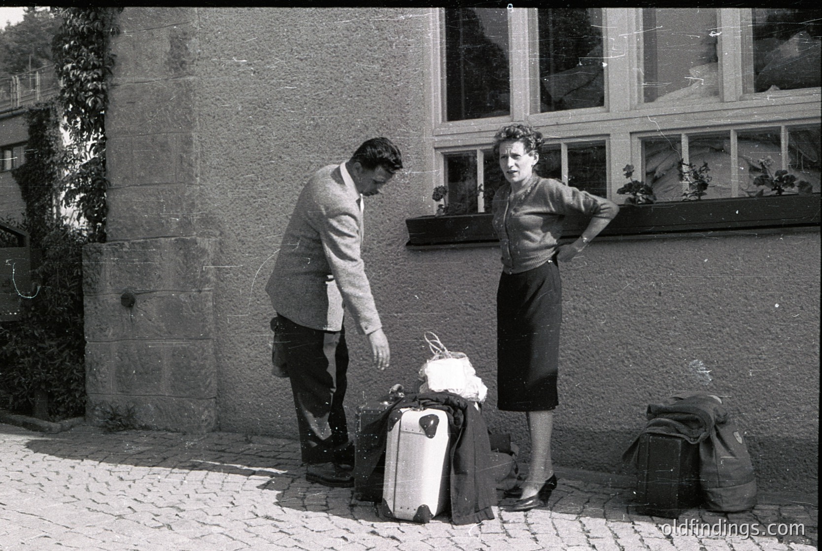 Mid-20th century street scene: man in suit and tie bends to secure a vintage suitcase, woman in structured dress leans against stone building. Decorative window boxes with plants flank a large rectangular window.