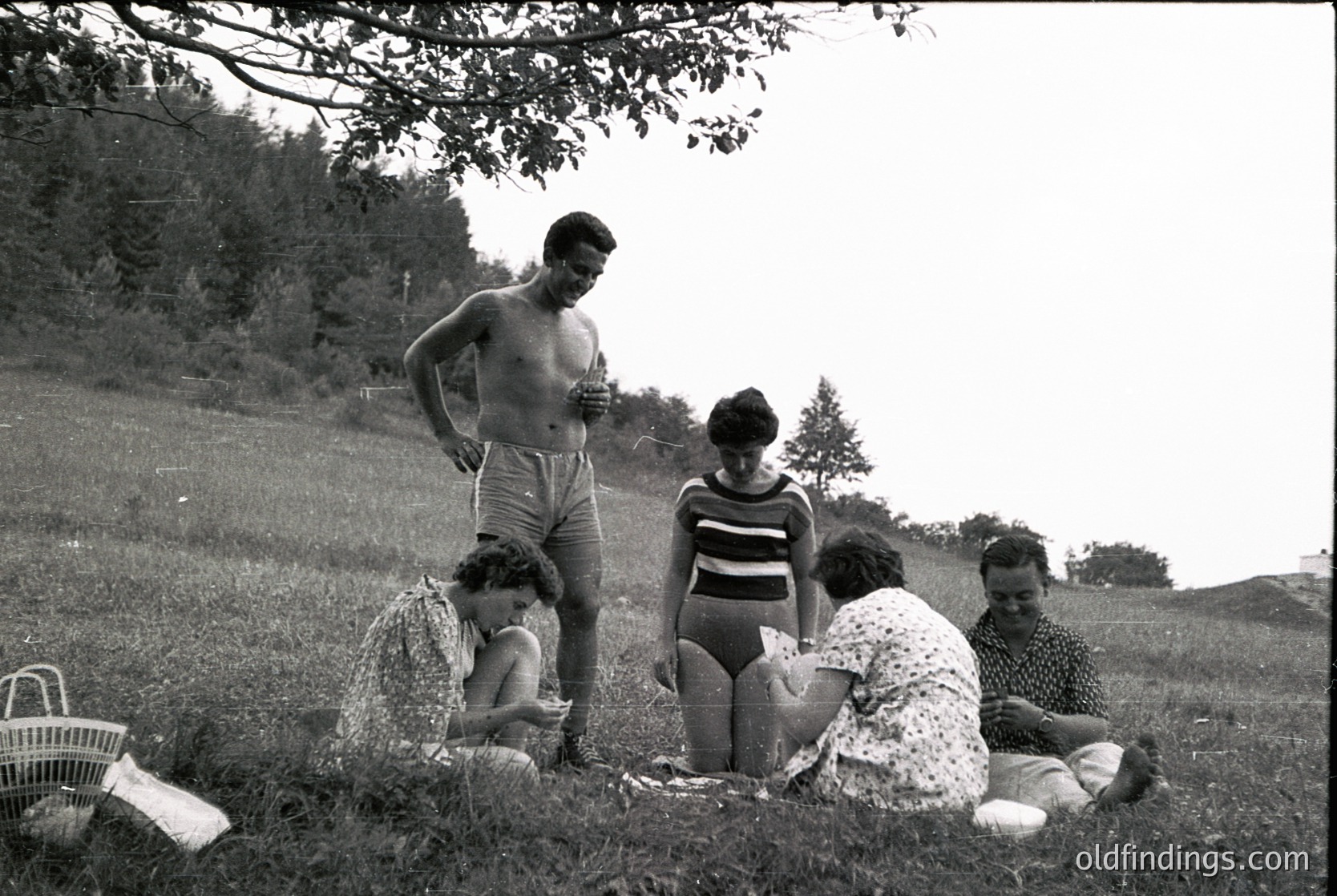Mid-20th century family picnic in a grassy field, likely 1950s–1960s. Five individuals—two adults standing, three seated—pose casually around a spread of food and a woven basket. The man stands shirtless in shorts, while others wear patterned swimwear and floral dresses. Lush greenery and trees frame the scene, suggesting a rural or suburban outdoor gathering.