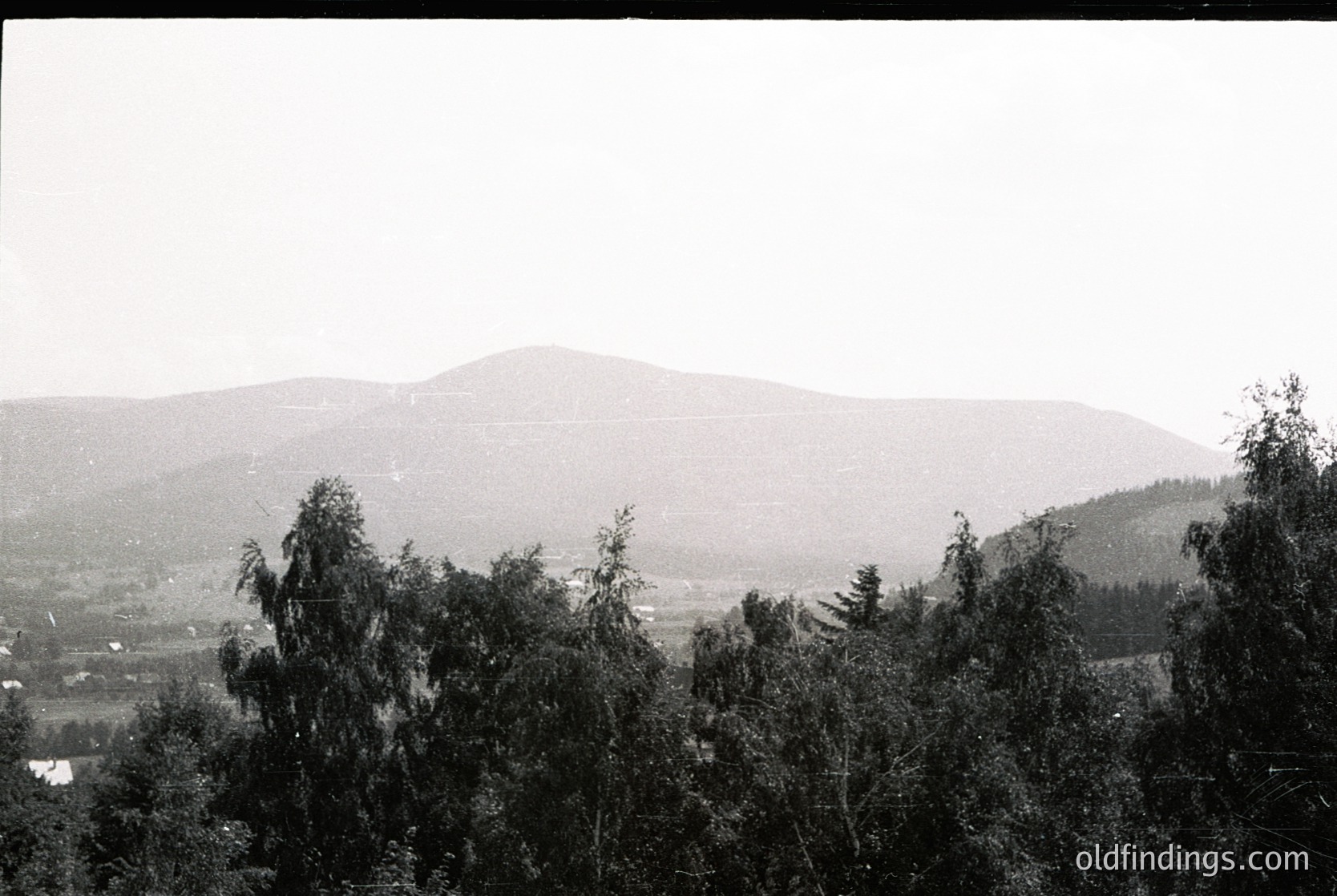 Vague black-and-white landscape featuring mist-covered rolling hills and sparse forest. Low-contrast composition with minimal human presence, likely mid-20th century.