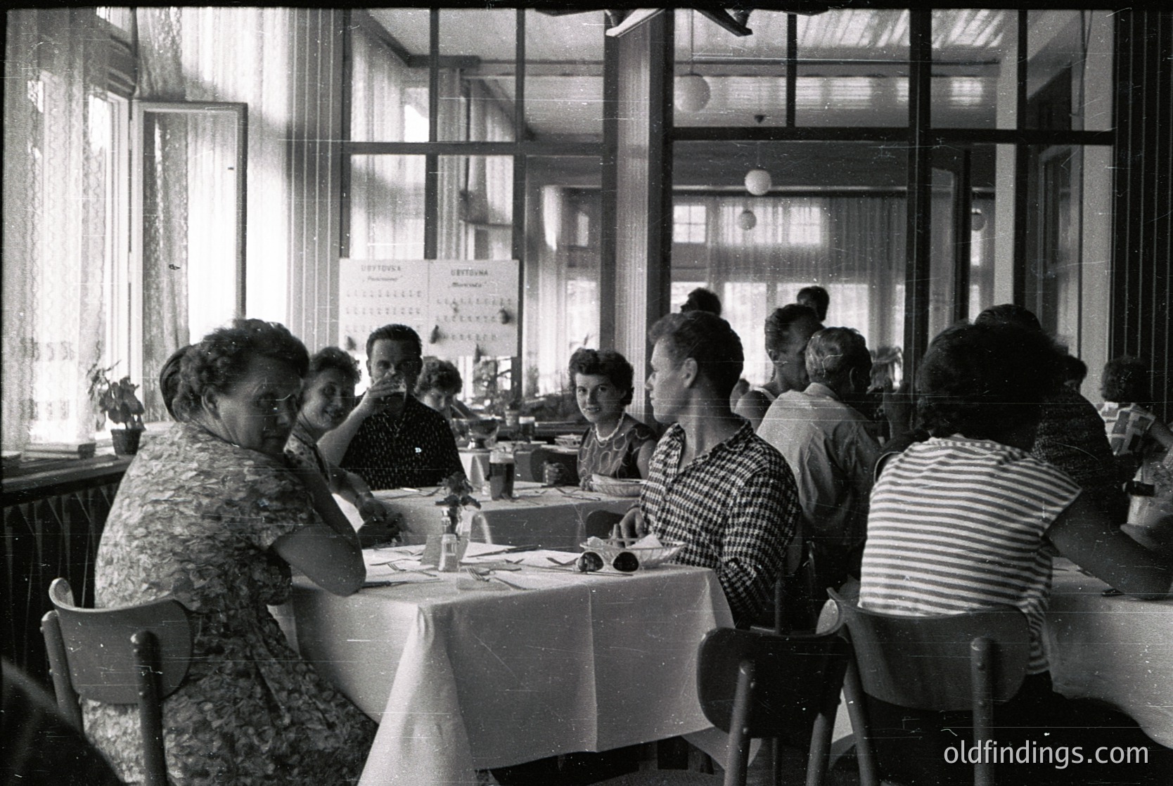 Mid-century café interior with 1950s-60s women dining at round tables, clad in floral and striped dresses. Natural light floods through large windows, revealing a cityscape beyond. White tablecloths, ashtrays, and vintage signage hint at a social, communal atmosphere. é