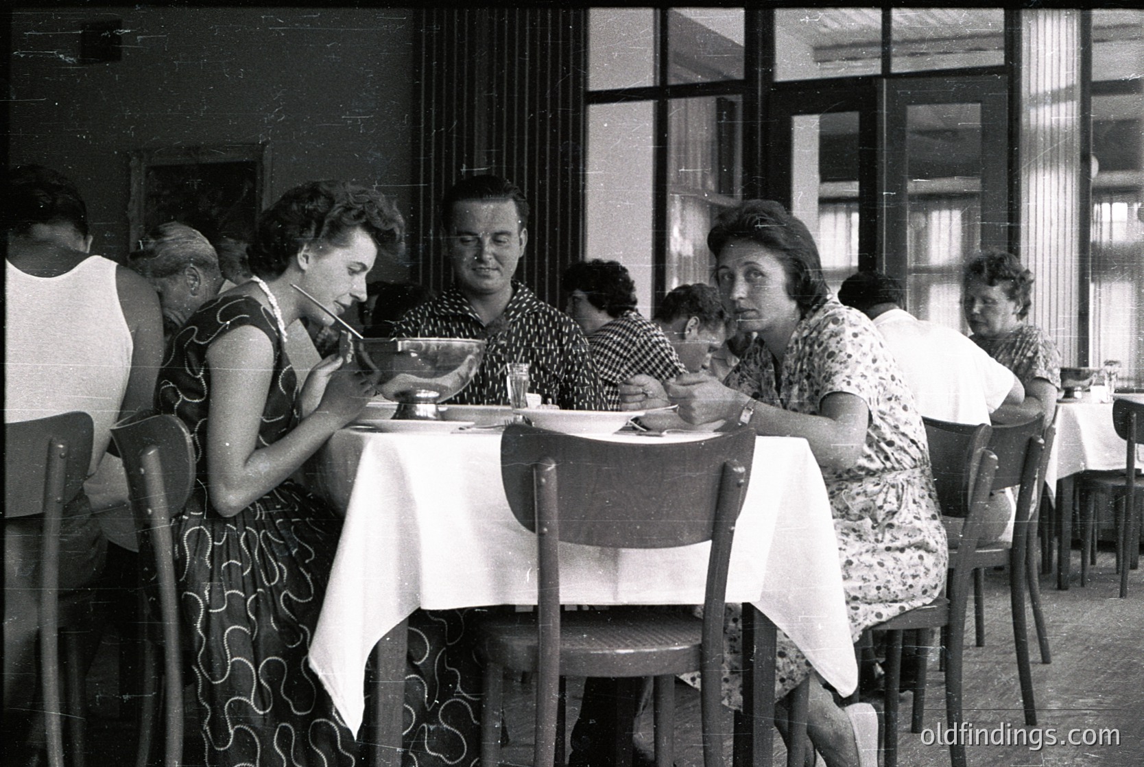 Mid-century café scene with six patrons seated at a long table, dressed in 1960s-70s attire. White tablecloths, metal chairs, and large windows with curtains suggest a European or Soviet-era dining hall. Men and women engage in conversation over plates and cups, reflecting social dining norms of the era. éCulture