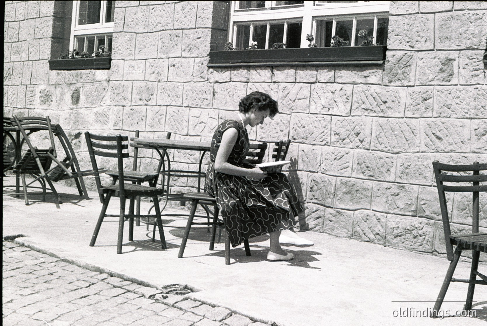Mid-20th century woman reading outdoors at a café table, dressed in a floral dress. Stone building with flower boxes in windows and cobblestone pavement. Empty wrought-iron chairs and tables suggest quiet afternoon. éCulture