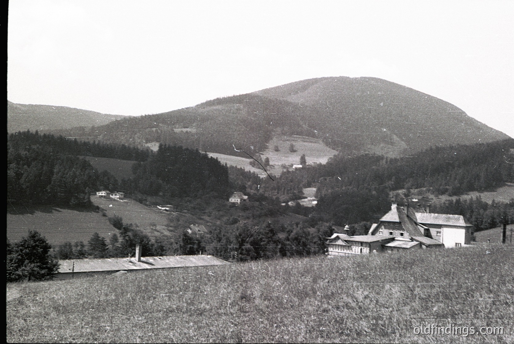Rural alpine valley with scattered farmhouses and dense forest. Mid-20th century architecture—sloped roofs, stone foundations—surrounded by rolling hills and open fields. Monochrome vintage aesthetic suggests early-to-mid European countryside.