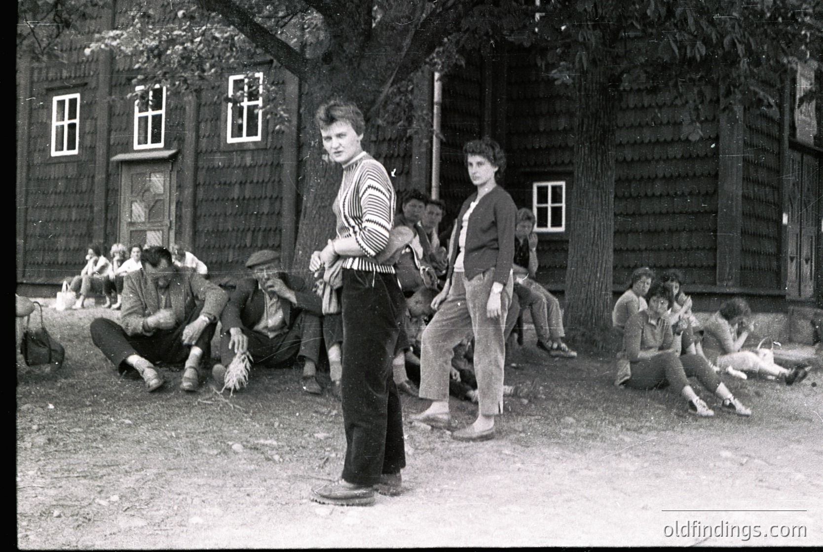 Black-and-white snapshot of a rural gathering, likely Eastern Europe, 1960s–1970s. Group of young people in casual attire—striped shirts, rolled-up jeans, and flat caps—pose and sit on a dirt path near rustic wooden buildings. One person stands prominently in the foreground, holding a cigarette. Others relax on hay bales or the ground, suggesting a communal, informal event.